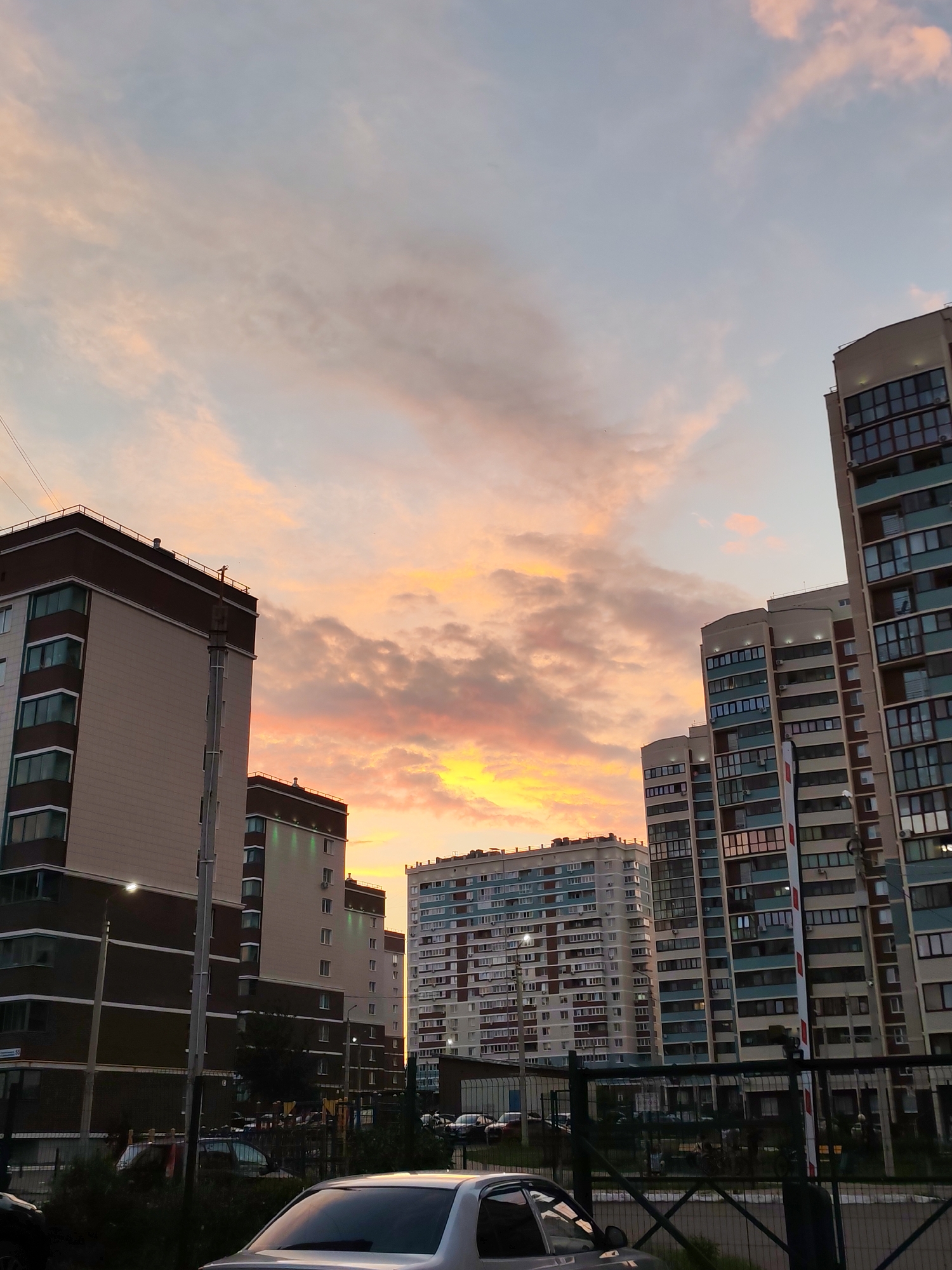 This image captures a sunset view in Izhevsk, Russia, dominated by tall residential buildings. The sky exhibits a gradient of colors, transitioning from a pale blue at the top to warm hues of orange and yellow near the horizon, with scattered clouds adding texture. In the foreground and midground, several multi-story apartment buildings are visible. One building on the left is a light beige color with dark brown accents, featuring rows of windows. To its right and further back, a cluster of taller buildings with a mix of beige, white, and teal facades are present. The building on the far right is particularly tall and has a grid-like pattern of windows. The lower parts of the image show the base of these buildings, including fences, streetlights, and the tops of parked cars. The scene appears to be from a residential district, likely taken from a slightly elevated position or across a street, looking towards a complex of modern apartment blocks during the twilight hours. No people are clearly discernible in the image.