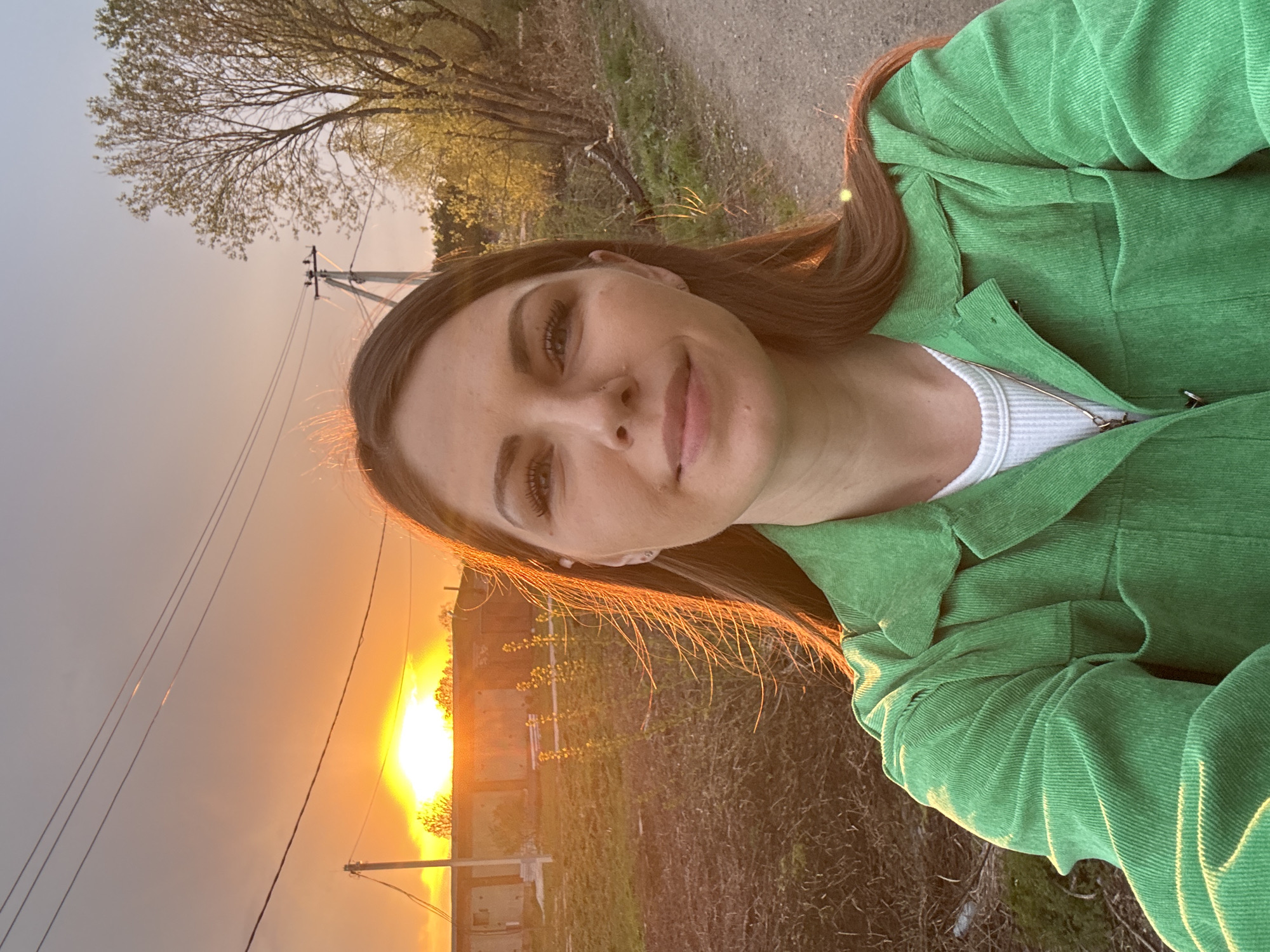 A woman is standing outdoors in Razdolye, Russia, during a sunset. She is wearing a green corduroy jacket over a white shirt. Power lines and utility poles are visible in the background, along with a building or structure. A tree is visible in the upper left corner. The ground is covered in vegetation.