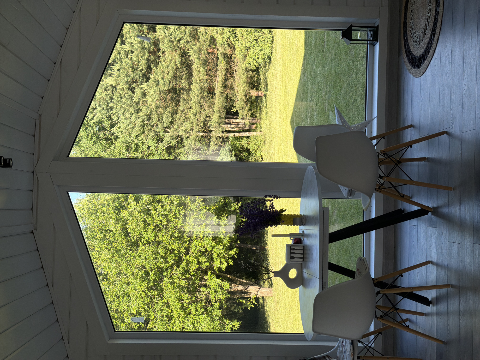 The image depicts a sunlit dining area inside a white-paneled room, with a large, triangular window offering a view of a lush green lawn and trees. A round, light-colored table with a dark, angular base stands in the foreground, set with three white chairs with wooden legs. On the table are decorative items including a white vase with a circular cutout, a rectangular holder with cutlery, a candle, and a yellow vase filled with purple flowers. To the right of the window, a black lantern sits on the floor next to a circular rug. The exterior scene shows a well-maintained lawn bordered by mature trees, suggesting a tranquil, possibly rural or suburban setting. There are no people present in the image. The lighting indicates it is daytime, with sunlight casting shadows on the floor and illuminating the foliage outside. The location is identified as Aleshkino DP, Russia.