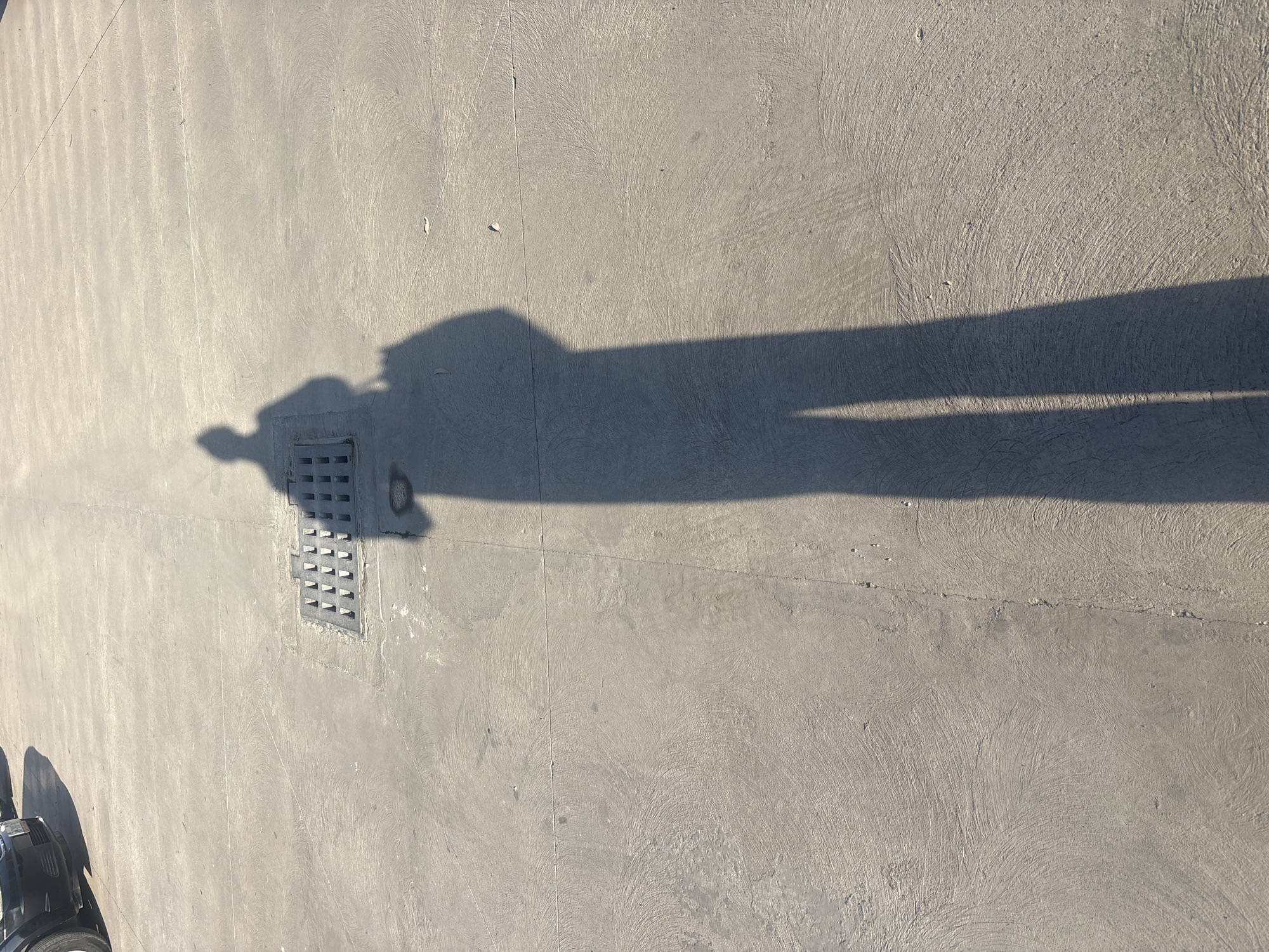 The image depicts a concrete surface outdoors, likely a sidewalk or paved area. A long, elongated shadow of a person is cast on the surface, stretching across the frame. A metal grate or drain cover is partially obscured by the shadow. A portion of a dark-colored object, possibly part of a vehicle or machinery, is visible in the lower left corner. Based on the presence of the grate and paved surface, the location is likely an urban or suburban area. Without additional information, it's impossible to confirm the specific location in Buenavista, Mexico.