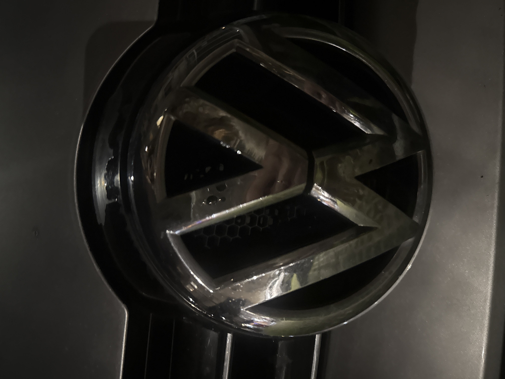 The image shows a close-up of a chrome Volkswagen emblem attached to a dark gray vehicle grille. The logo is a circle containing a stylized "VW" symbol. The background behind the logo is a honeycomb pattern. There is no information about the location being in Varna, Bulgaria, or any people, actions, interactions, or environmental context visible in the image.