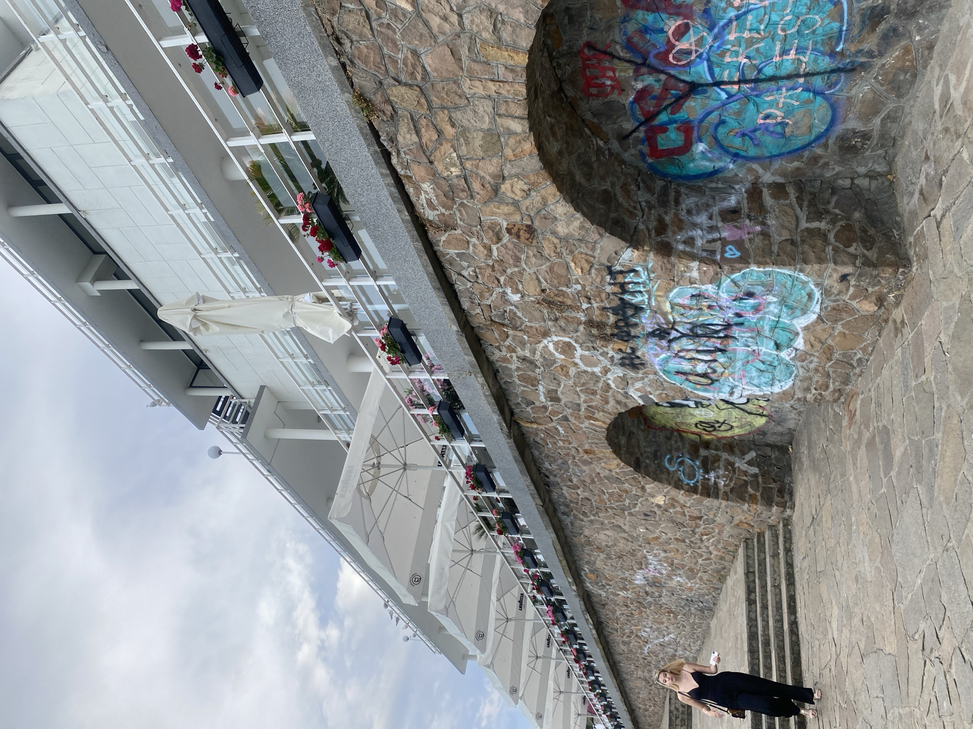 An eye-level, full shot captures a young woman walking down a stone staircase towards the viewer on a cloudy day. She is dressed in a black jumpsuit and sandals, carrying a white object in her right hand and a purse on her left shoulder. Behind her, a textured stone wall with arched openings covered in graffiti dominates the foreground. Above the wall, a white building with multiple balconies, white railings, and rows of potted red and pink flowers stretches across the frame. Several large, white umbrellas are extended from the building's balconies, suggesting an outdoor dining or seating area. Seagulls are perched on the railings of the building. The scene is located in Burgas, Bulgaria, likely along the Sea Garden promenade, known for its artistic displays and connection to the sea.
