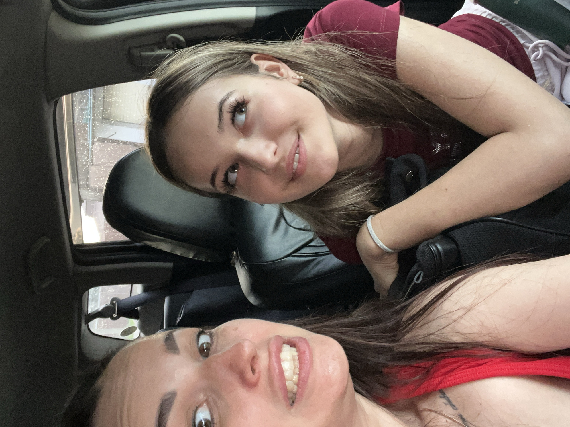 Two young women are in the passenger seats of a car. The woman in the foreground on the left is looking at the camera with her mouth open, revealing her teeth. She has dark brown hair and is wearing a red top. The woman on the right is looking upwards and to the right, with a slight smile. She has blonde hair and is wearing a maroon top. The interior of the car is visible, including the headrests of the seats. The background appears to be a view through the car windows, showing trees and buildings.