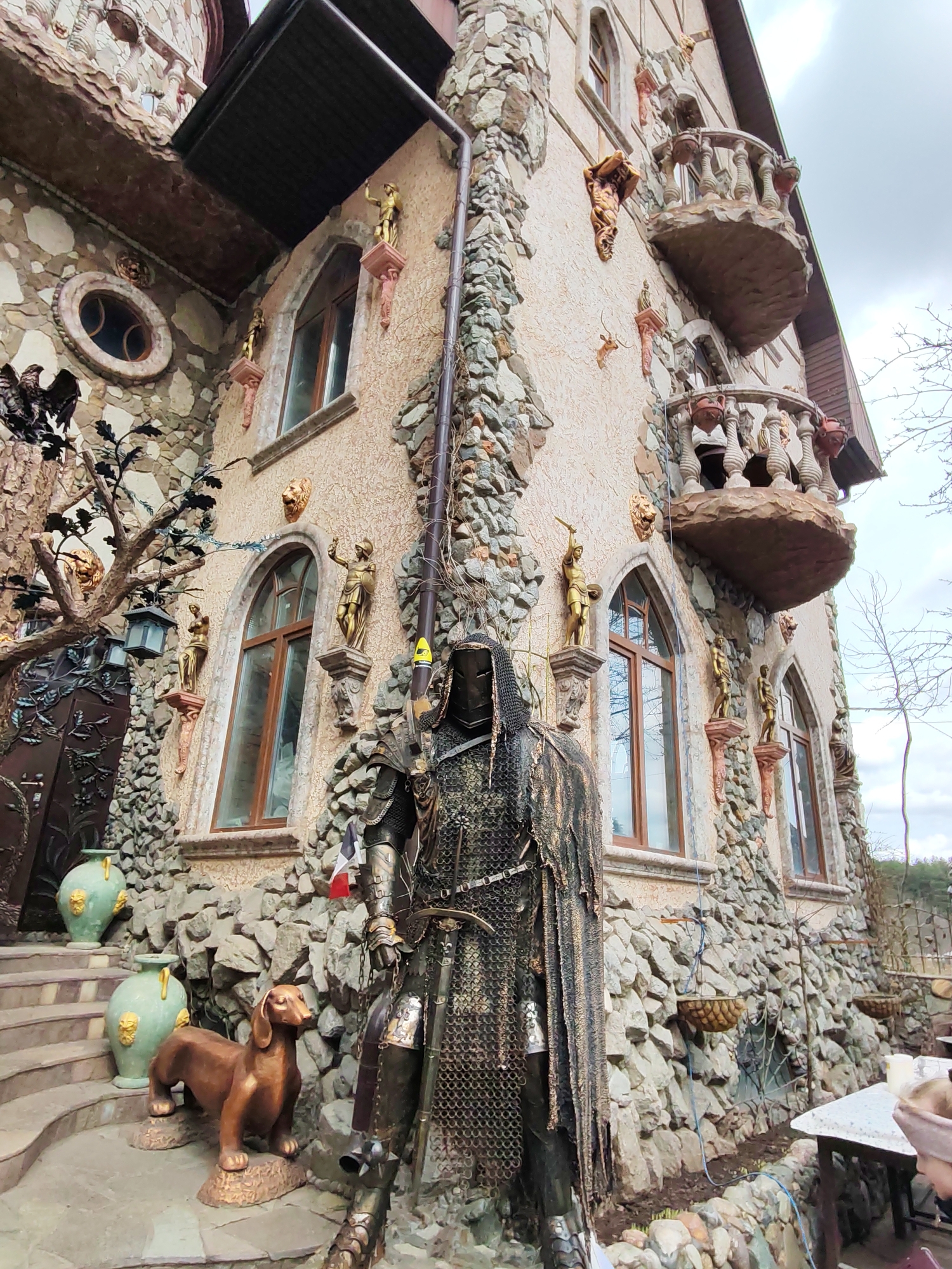 The image shows a whimsical building in Izhevsk, Russia, constructed of stone and decorated with sculptures and ornate details. In the foreground stands a metal knight statue, draped in chainmail. To the left, a set of stairs leads to an arched doorway, flanked by two pale green vases. On the steps is a brown dachshund statue. The building's façade includes arched windows, balconies with figures, and numerous golden statues mounted on pedestals. The building is surrounded by a stone wall, and a table is visible on the right side of the frame. The sky is overcast.