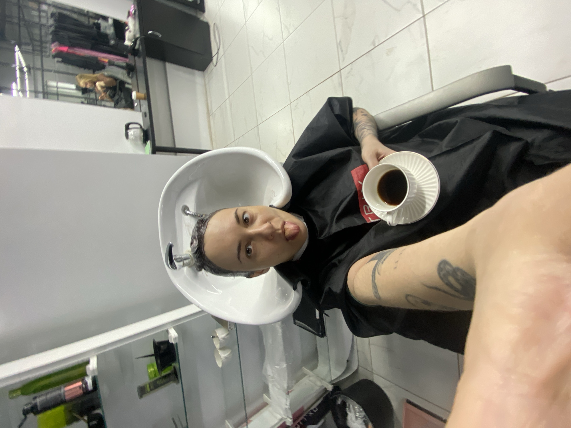 A person lies back in a salon chair with their head in the washbasin. They are wearing a black salon cape and have wet hair. The person holds a cup of coffee in one hand and is sticking their tongue out at the camera. The location appears to be the interior of a hair salon. It is not possible to determine the exact location in Votkinsk, Russia, based on the image alone.