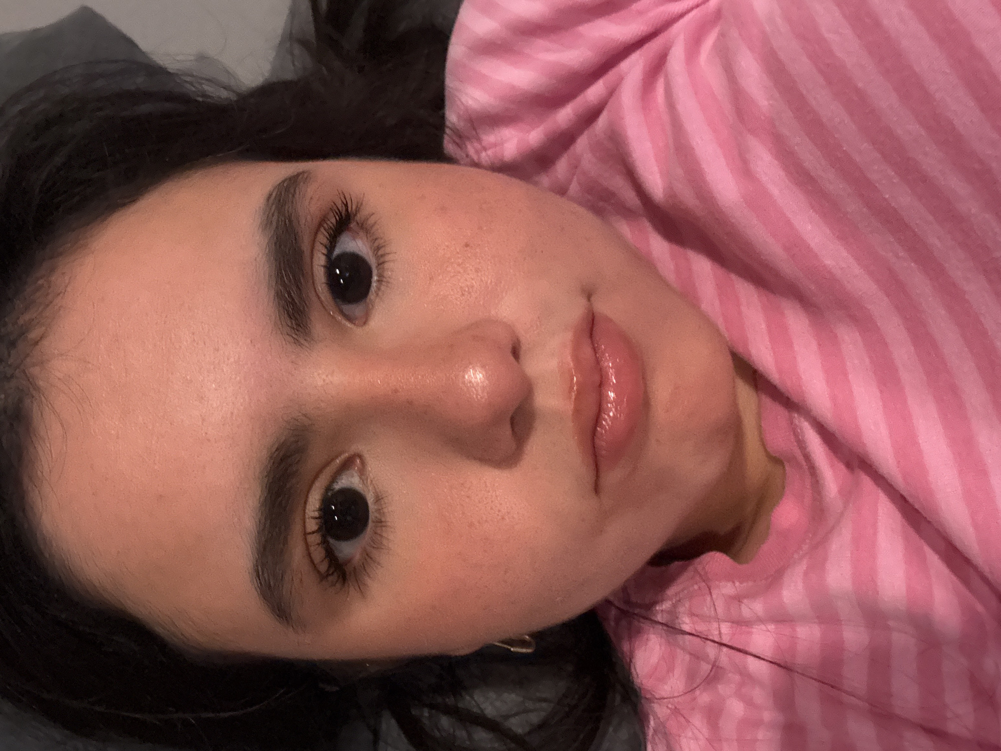 A close-up, frontal medium shot captures a young person's face, with dark hair framing their features. They are wearing a pink and white striped t-shirt. The person's skin appears flushed, with subtle speckles of freckles visible. Their eyebrows are dark and full, and their eyelashes are long and dark. The eyes are open and dark, reflecting light. The nose is slightly shiny, and the lips are glossy, with a slight pout. The background is a dark, indistinct surface. The scene is set in Austin, Texas, United States of America.