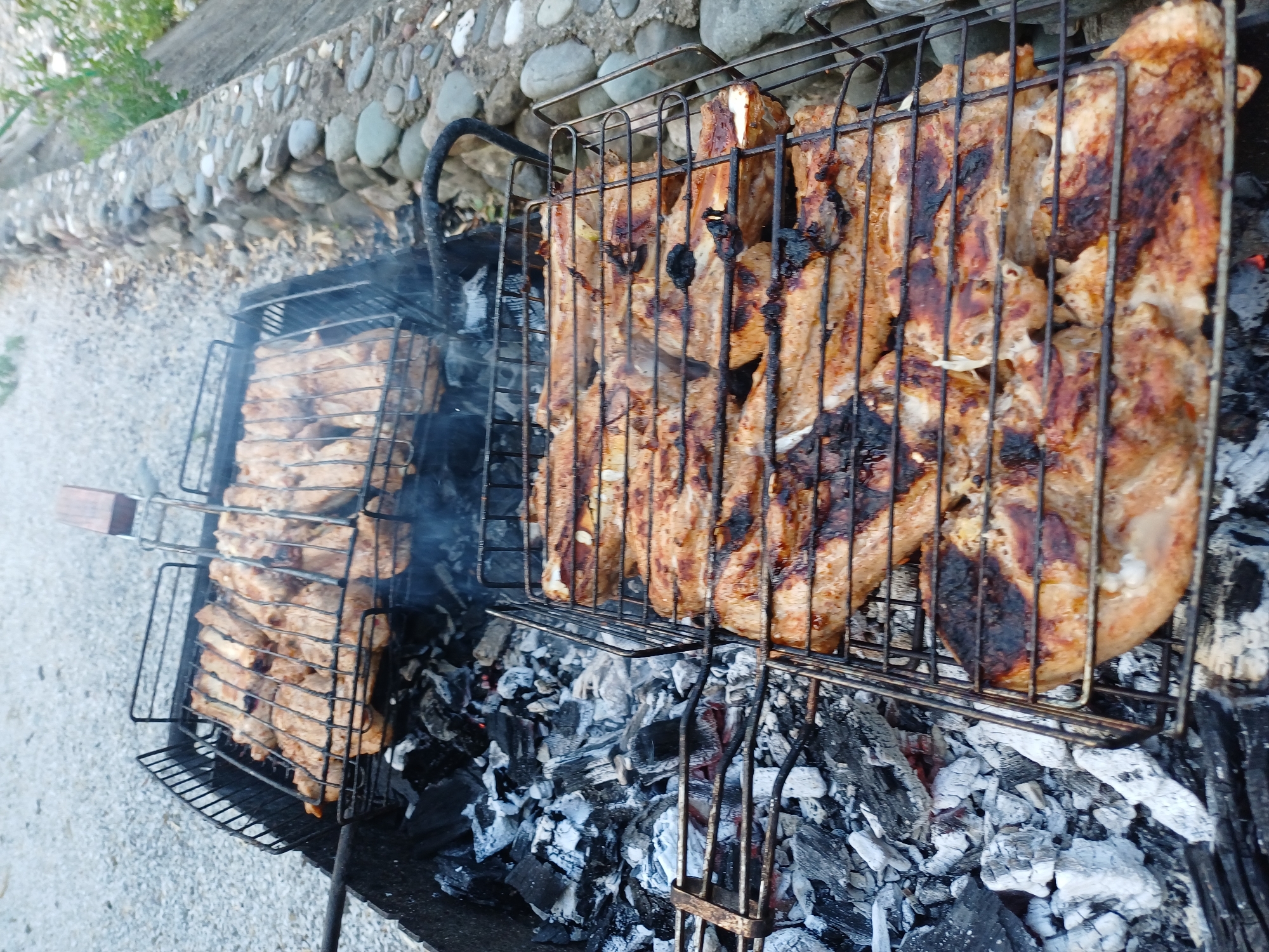 A high-angle shot captures meat grilling over charcoal in what appears to be an outdoor setting, likely in Monastyr, Russia. The meat, consisting of numerous ribs or chops, is arranged on two metal grill baskets placed over a bed of glowing charcoal and ash. The meat on the lower basket is more browned and shows some charring, while the meat on the upper basket is lighter in color. Smoke rises from the grill, indicating active cooking.
The environment surrounding the grill consists of a gravel-covered ground and a stone wall made of rounded river stones. Some sparse greenery is visible behind the stone wall. The overall scene suggests a casual outdoor meal preparation. There are no people visible in the image.