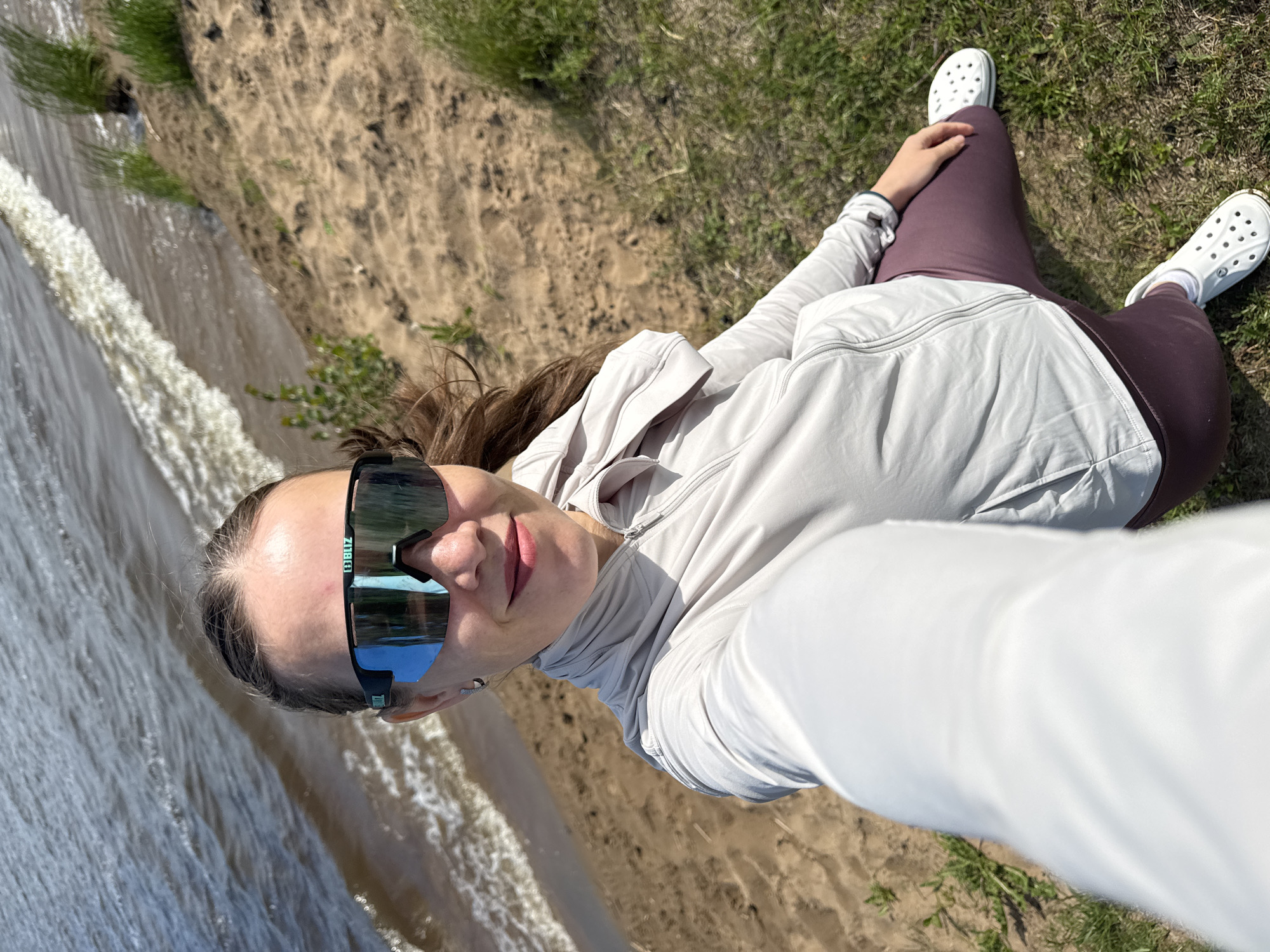The image features a woman taking a selfie outdoors, positioned on what appears to be a sandy or dirt bank next to a body of water, consistent with a river or reservoir in Chaikovsky, Russia. The perspective is a high-angle shot, looking down at the subject and her immediate surroundings.
The subject is a woman with long, dark brown hair, wearing a light gray or beige full-zip hooded jacket, dark maroon leggings, white socks, and white Crocs shoes. She is wearing large, reflective sports sunglasses, with the brand name "BLIZ" visible on the right arm of the frame. Her expression is neutral to subtly smiling.
The scene includes a body of water, possibly a river or reservoir, which takes up the left side of the frame. The water is choppy with visible ripples and some white foam near the bank. To the right of the woman, the ground is sandy or dirt-covered, transitioning into patches of green grass further back.
The activity taking place is the woman posing for a selfie. The lighting suggests a bright, sunny day, likely during daylight hours. No specific time of day is indicated beyond daylight. The setting is clearly outdoors and natural.