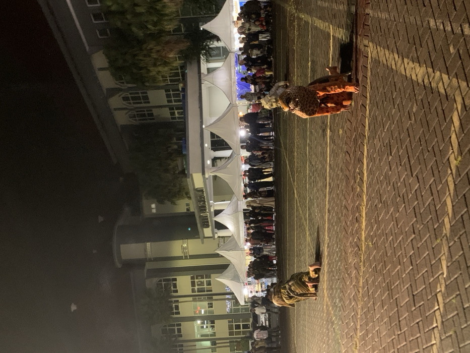 The image shows a night scene in front of a building in Kecamatan Batu, Indonesia, located at latitude -7.867 and longitude 112.514. There are numerous white tents in the foreground, lined with spectators. Two people are in the foreground, facing away from the camera, dressed in animal costumes, kneeling on the brick pavement. The pavement appears to be a parking lot or walkway. The scene is brightly lit. The building behind the tents is a two-story structure with lit windows. Palm trees are visible in the distance.