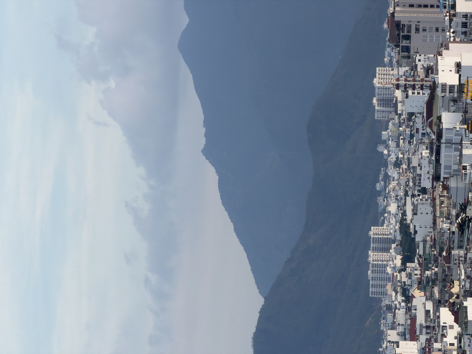 The image shows a panoramic view of the city of Nha Trang, Vietnam, with mountains in the background. The city is densely populated with a mix of low-rise buildings and several high-rise apartment buildings. The architecture appears to be a mix of modern and traditional styles, with many buildings painted white. The sky is overcast with clouds. In the foreground, there are several buildings with red tile roofs. A large white apartment building stands out in the middle ground. The mountains in the background are covered in vegetation and appear to be part of a mountain range. The overall impression is one of a bustling city nestled between the mountains and the sea.