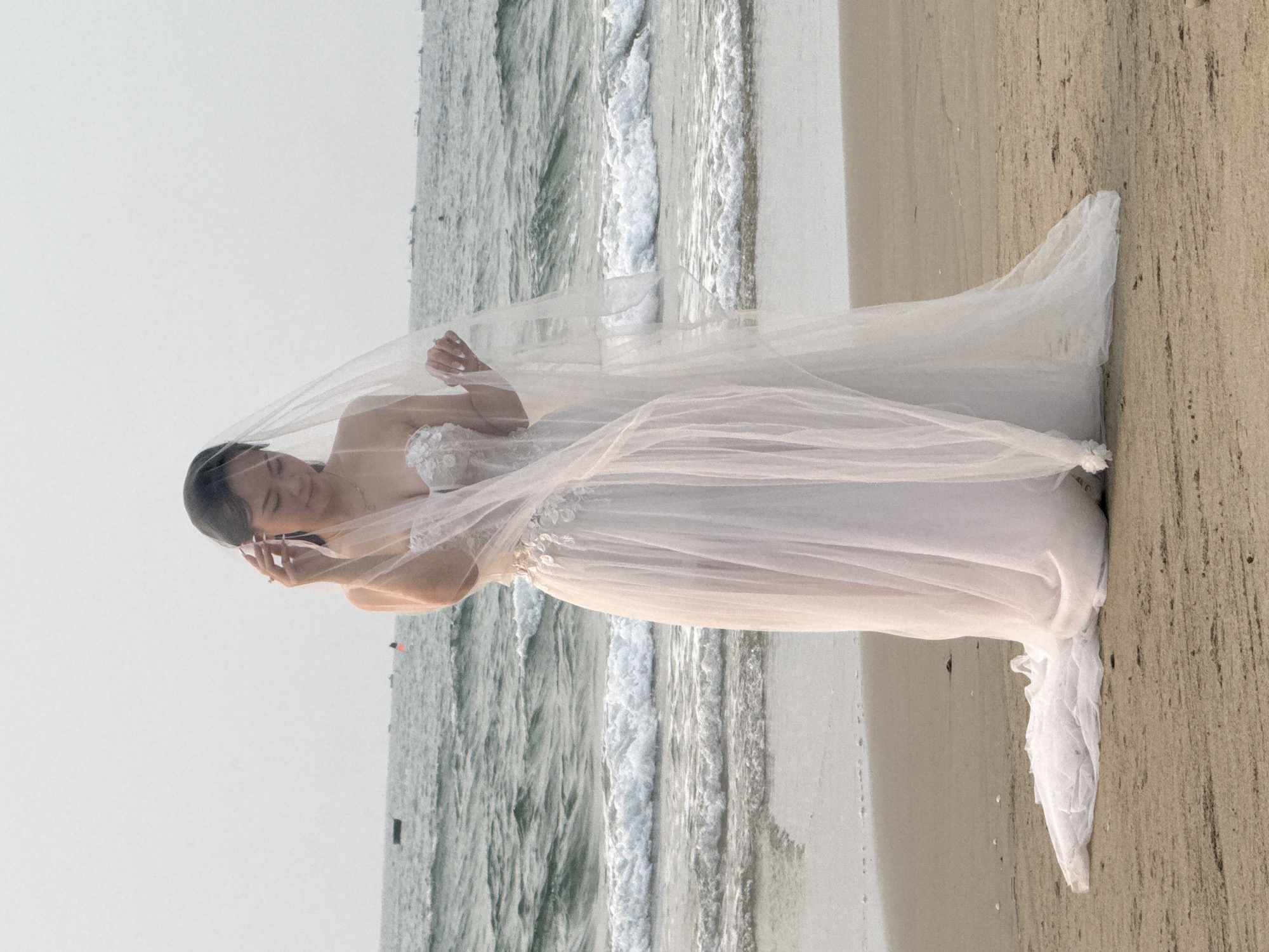 A bride in a white wedding dress and veil is standing on a beach in Đà Nẵng, Vietnam. The waves are crashing behind her, and the beach stretches out in both directions. The bride is wearing a white wedding dress with floral embellishments. The veil is light and airy, and it partially covers her face. She is smiling, and her eyes are closed. She holds the veil in front of her face with her hands. The beach is a light brown color, and the water is a light blue color. The sky is overcast.