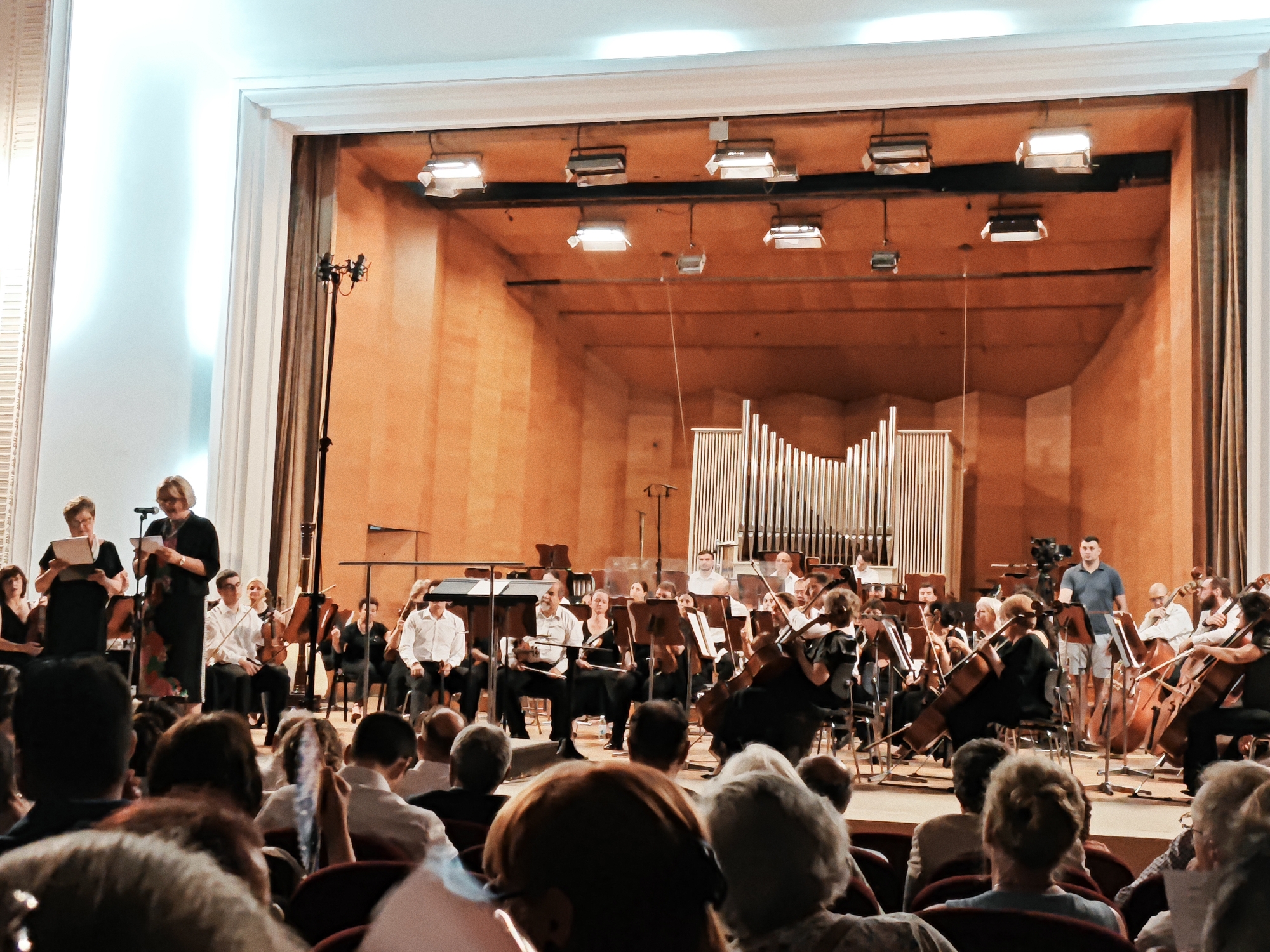 This media file depicts an indoor orchestral performance. The scene is set on a stage with warm, wooden paneling and an organ in the background. A large orchestra is assembled, with musicians playing various instruments including cellos, violins, and possibly a piano. Several conductors or soloists are visible, some standing and addressing the audience or directing the orchestra. The audience is seated in the foreground, facing the stage, with some individuals holding programs or sheet music. The lighting appears to be stage lighting, focused on the performers. The atmosphere suggests a formal or semi-formal event, likely a concert or recital. There is no visible text within the frame.