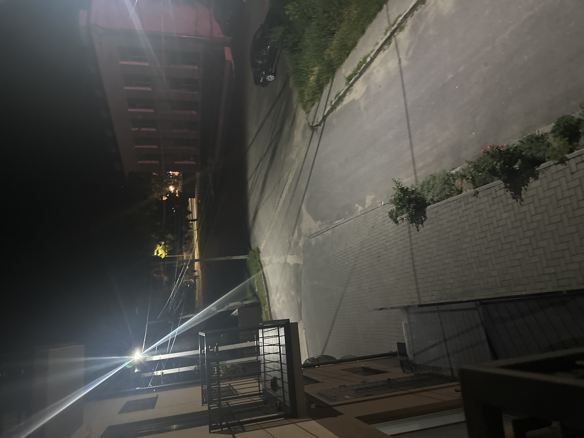 An outdoor, night-time, high-angle view captures a street scene with buildings and a paved walkway in Strășeni, Moldova. The foreground features a partial view of a building with a metal-railed balcony and a paved walkway with a brick pattern. To the right, a road is visible with tire marks and a dark-colored car parked along the side. A building with multiple windows, appearing to be a two-story structure, is situated across the street. The lighting suggests artificial illumination from streetlights, casting long shadows across the scene. Vegetation, including bushes and grass, lines the edge of the road. In the distance, a few indistinct figures can be seen walking on the street. The overall atmosphere is one of a quiet urban environment at night.