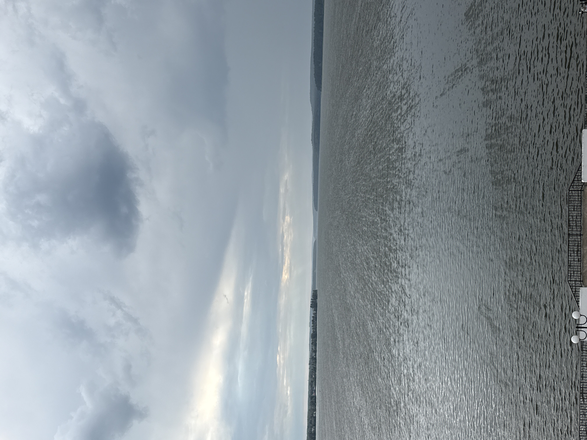 The image shows a wide, dark body of water under a cloudy sky. The water has small ripples, creating a textured surface. In the distance, a narrow strip of land is visible, with trees and some buildings suggesting a shoreline settlement. The sky is dominated by heavy, grey clouds, with some brighter patches where the sun is attempting to break through, casting a diffused light. In the foreground, a black metal railing runs along the edge of a paved area, with a white structure and two lamp-like fixtures visible. This scene is likely a waterfront in the city of Votkinsk, Russia, possibly overlooking the Votkinsk Reservoir on the Kama River.