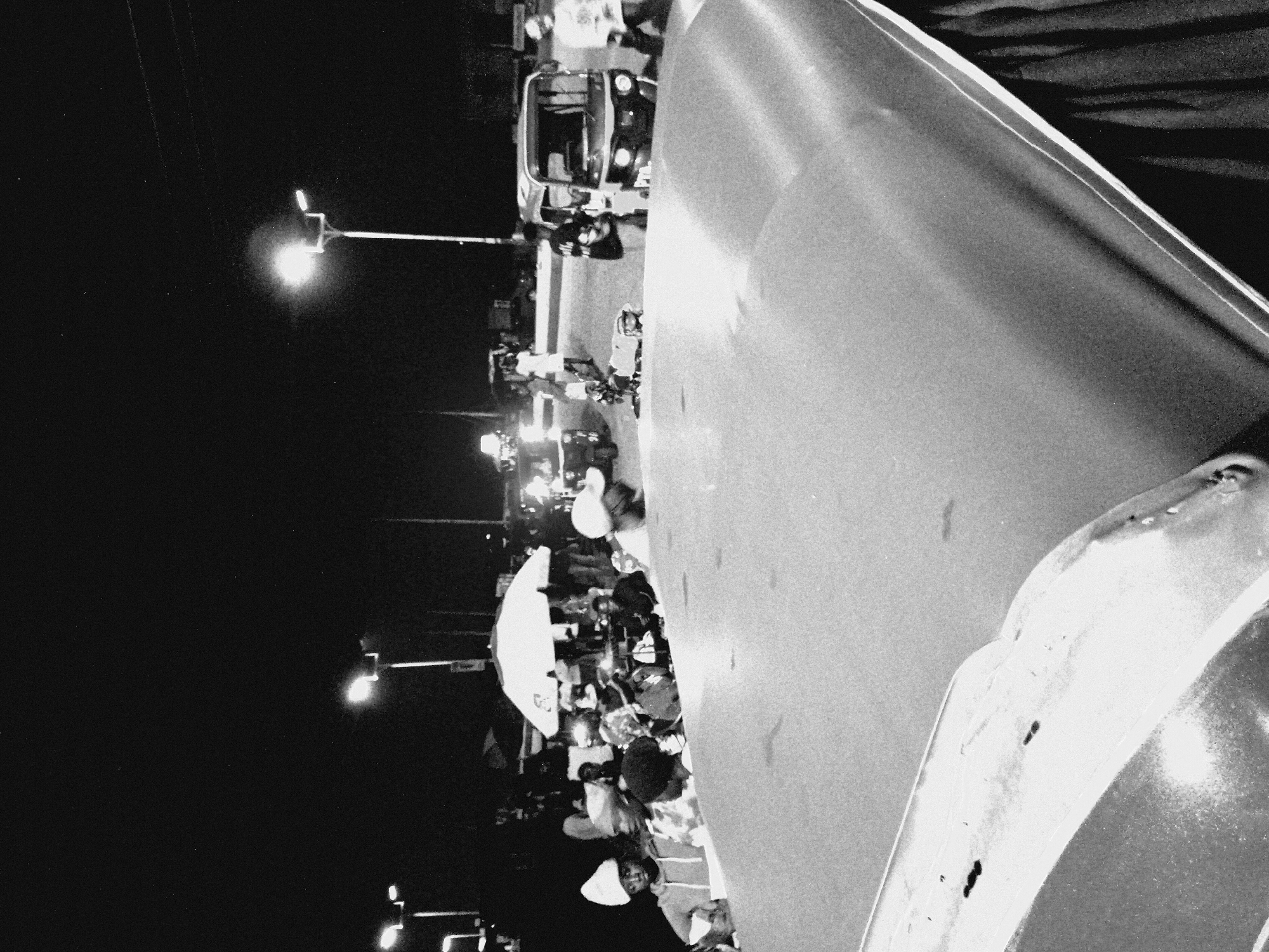 The image depicts a nighttime scene in Bukuru, Nigeria. In the foreground, the curved, metallic surface of what appears to be the hood of a vehicle is prominently featured, with a dark, fabric-like material visible along the right edge. Beyond this, a group of people are gathered. Some individuals are wearing hats, including a white beanie on a person in the lower left. In the mid-ground, an open white umbrella is visible, and several people are standing or sitting around it, possibly at a market stall or gathering. Behind the people, several tuk-tuks (auto rickshaws) are present. One tuk-tuk is in the center-right of the image, and another is visible to its left. Streetlights illuminate the scene, with a bright streetlight positioned in the upper right quadrant of the image. The background is dark, with the sky and distant, indistinct structures. The overall impression is of a busy, dimly lit urban environment at night.