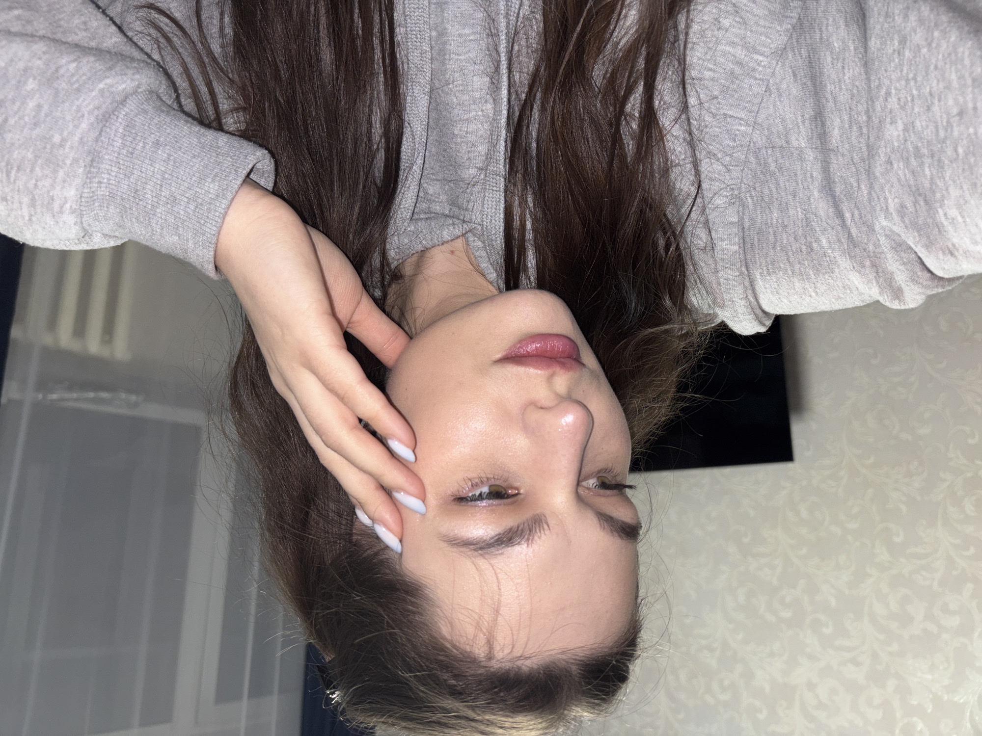 A woman with brown hair and fair skin is wearing a gray hooded sweatshirt. She is holding her hand near her face. The background consists of a wall with a floral pattern, a window, and a television. The image is likely taken indoors. The image is upside down. There is no information to verify the location as Chaikovsky, Russia.