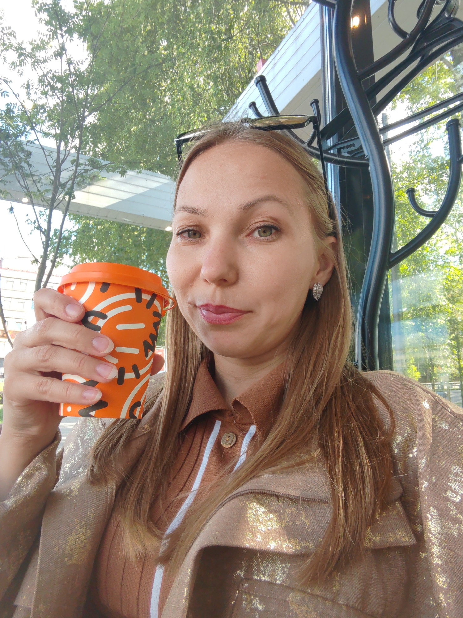 A woman with blonde hair is holding an orange paper cup with a lid, likely containing a beverage, in her left hand. She is wearing a light brown, collared shirt with a vertical white stripe and a button closure. Over the shirt, she has a shimmering beige jacket with a textured pattern. A pair of sunglasses is perched on top of her head. She is wearing a decorative silver earring in her visible right ear. She appears to be looking directly at the camera, with her mouth slightly open. The background shows a blurred view of greenery and trees through a window, suggesting an outdoor or semi-outdoor setting, possibly a cafe or restaurant. The overall lighting suggests it is daytime. The image's metadata does not contain information about the specific location in Syktyvkar, Russia.