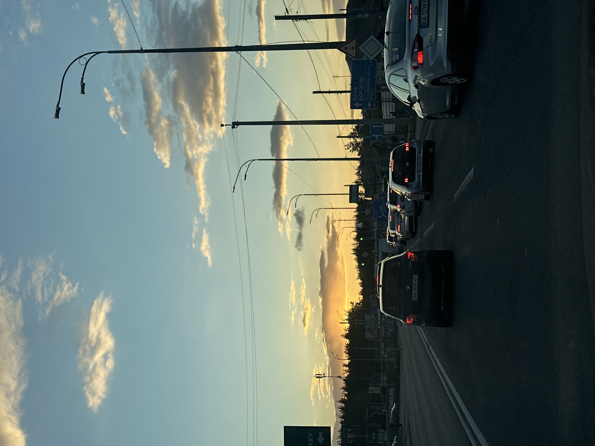 The image, rotated 90 degrees clockwise from its original orientation, displays a road scene at what appears to be sunset or sunrise. The sky, occupying the upper portion of the frame, transitions from a clear light blue overhead to warm yellow and orange hues near the horizon, illuminated by the low sun. Scattered, elongated clouds, catching the golden light, are visible across the sky.
A multi-lane asphalt road with white markings occupies the lower part of the scene. Several cars are visible, moving in a line, suggesting traffic. Their rear lights are prominent. Along the side of the road, a series of tall utility poles or streetlights with curved arms extend upwards. To the right, a dark silhouette of trees forms a clear horizon against the bright sky.
Prominently displayed road signs provide location context. One sign points towards "БУЧА," "ВОРЗЕЛЬ," and "ІРПІНЬ." Another sign indicates "ГОСТОМЕЛЬ." These are all towns in Ukraine. Ukrainian license plates, such as "KA 3298 E" and "AA 3674 HT," are visible on the vehicles, confirming the setting. The scene captures the end or beginning of a day on a busy road in a suburban or inter-city area of Ukraine.