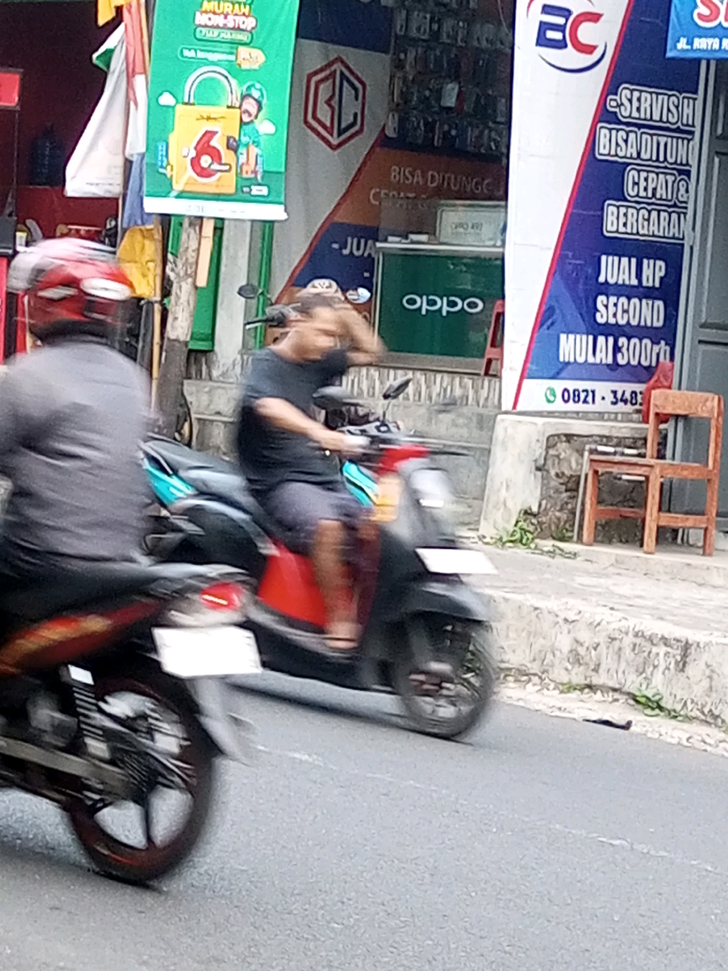 The image depicts a street scene in Mojotengah, Indonesia, featuring two individuals on motorcycles in motion, suggesting a commercial area. In the foreground, a person wearing a grey shirt and red helmet is on a motorcycle, blurred due to movement. Behind them, another person in a black t-shirt and dark shorts is also on a motorcycle, with their right hand raised towards their head.
The background reveals several commercial banners and signs. One prominent green banner features a yellow padlock with the number "6" and the text "MURAH NON-STOP". To the right, a blue and white banner displays text including "SERVISH HP", "BISA DITUNG", "CEPAT & BERGARAN", "JUAL HP SECOND MULAI 300rb", and a phone number "0821 - 3483". Another sign with a red hexagonal logo containing "GC" is visible. A green display stand with the "oppo" logo is also present. The street is paved with asphalt, and a concrete curb runs along the side, with some vegetation growing in crevices. A wooden chair is placed on the curb. The overall lighting suggests daytime.