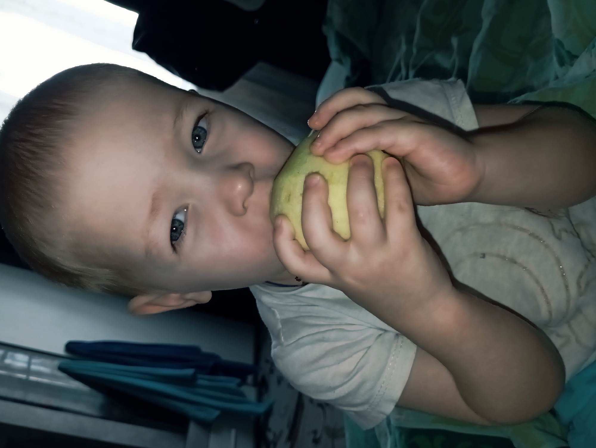 A young child with fair skin and blue eyes is holding and biting into a yellow fruit, possibly an apple or pear. The child is wearing a light-colored t-shirt. The background includes dark, undefined objects and a surface with blue and green cloth. The photograph appears to be taken indoors in an unknown location. The location of Yurgamysh, Russia is not ascertainable from visual inspection.