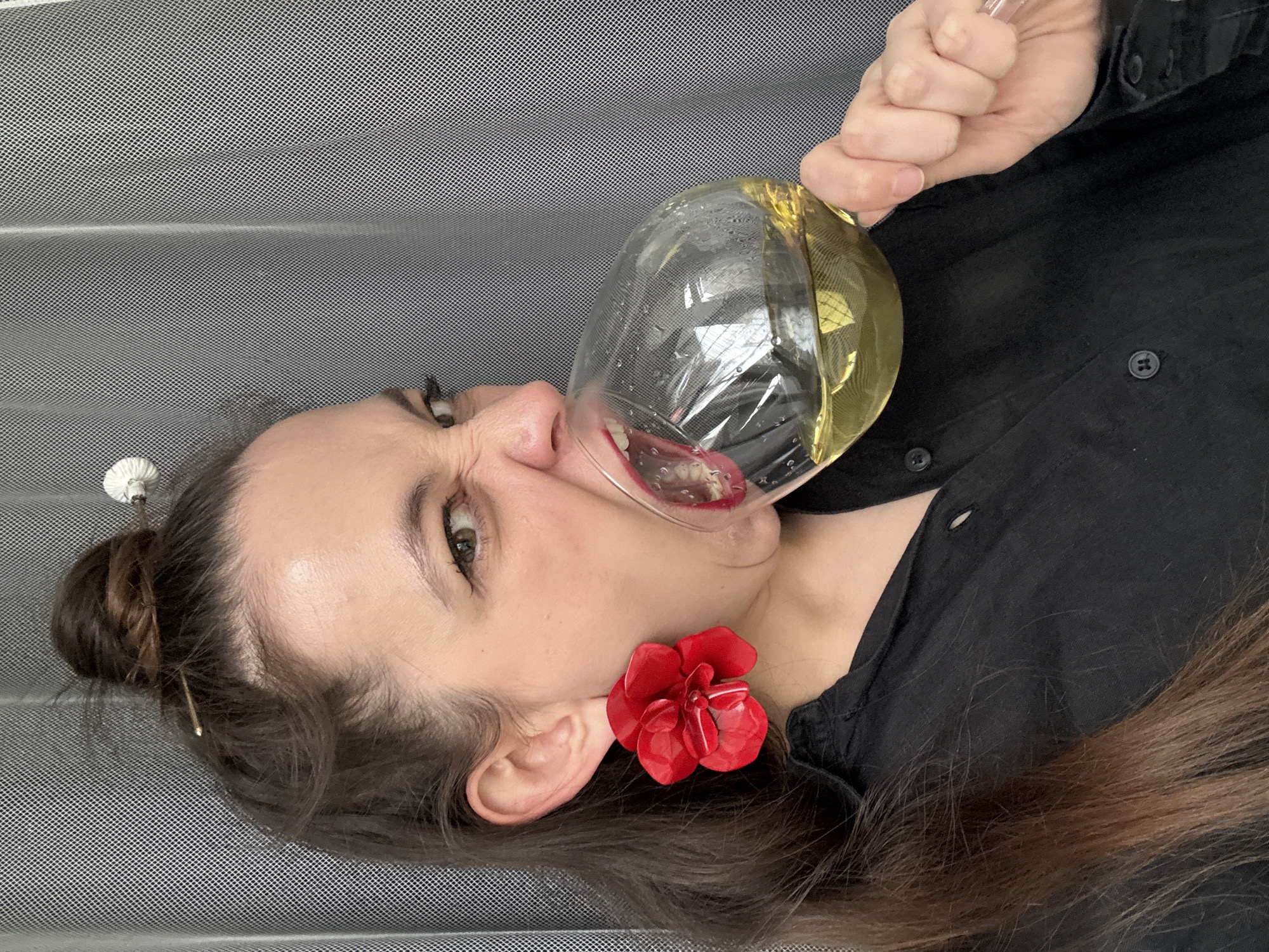 A woman is drinking from an oversized wine glass. She is holding the glass in her right hand, tilted so that the liquid inside is reaching her mouth. The woman is wearing a black button-up shirt and a large red flower earring on her left ear. Her hair is pulled back in a bun with a decorative pin. The background appears to be a grey patterned material. Based on prior knowledge, it can be inferred that the location is Chaikovsky, Russia, though this is not visually apparent in the image.