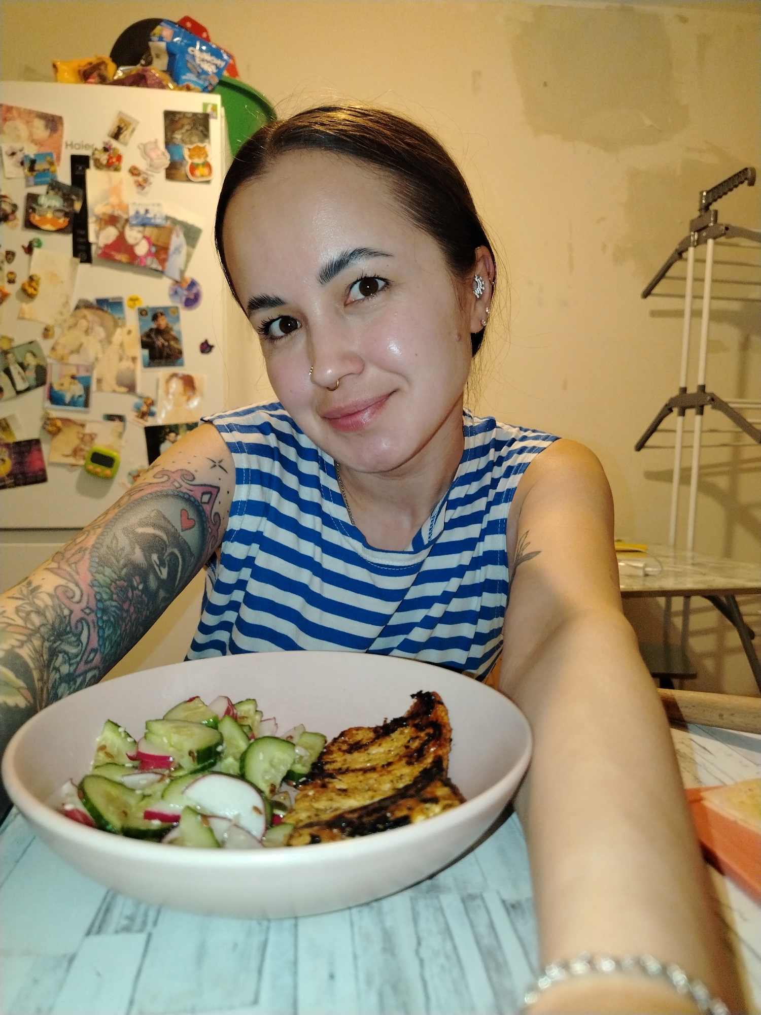 A young woman is captured in a close-up selfie, holding a light pink ceramic bowl of food in the foreground. She has dark hair pulled back, warm brown eyes, and a slight, pleasant smile, looking directly at the camera. She has a nose ring and a prominent snowflake-design earring in her right ear. Her left arm, closer to the camera, is covered in a colorful, intricate full-sleeve tattoo depicting various motifs including a badger-like creature, geometric patterns, and a small red heart. A simpler, leaf-like tattoo is visible on her right bicep. She is wearing a blue and white striped sleeveless top and a silver-colored bracelet on her right wrist.
The bowl contains a meal consisting of a fresh salad with sliced cucumbers and radishes, lightly sprinkled with seeds, alongside a piece of grilled food that shows distinct char marks, possibly chicken or fish. The table surface beneath the bowl has a light-colored, wood-grain pattern.
The background suggests an indoor, domestic setting in Noviy, Russia. To the left, a white "Haier" refrigerator is heavily adorned with numerous magnets, printed photos, and papers, creating a personal and lived-in feel. Various items, including a black cap, a green bowl, and snack bags, are stacked on top of the refrigerator. The wall behind her and to the right is a warm, light beige or yellowish color, appearing somewhat unfinished or marked in sections. A utilitarian white and gray clothes drying rack is partially visible on the right, next to another table. The overall lighting is warm, indicating artificial indoor illumination. The woman's expression conveys contentment as she appears to be enjoying or about to enjoy her meal.