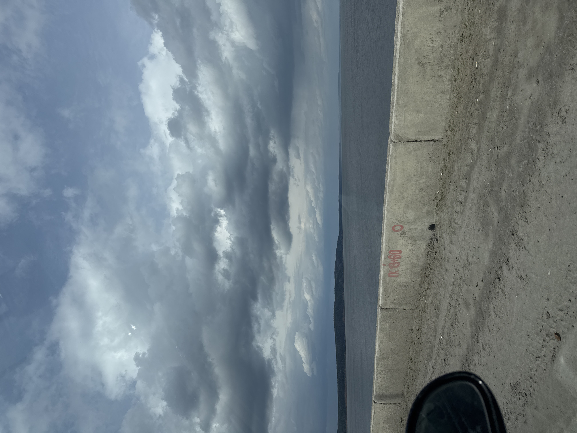 The image shows a cloudy sky, a body of water, and a concrete barrier. The concrete barrier has red markings that appear to be alphanumeric. Part of a side-view mirror is visible in the lower right corner. Based on the water body and the concrete barrier, the location is likely a bridge or embankment in Chaikovsky, Russia.