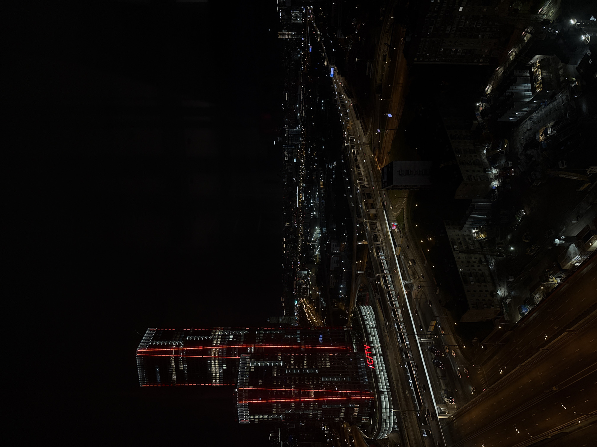 Aerial night shot of Khimki, Russia. A large, well-lit complex labeled "ICITY" is positioned near a multi-lane highway. Further in the distance is a city skyline lit with various points of light. A dark, unlit area occupies the left portion of the frame.