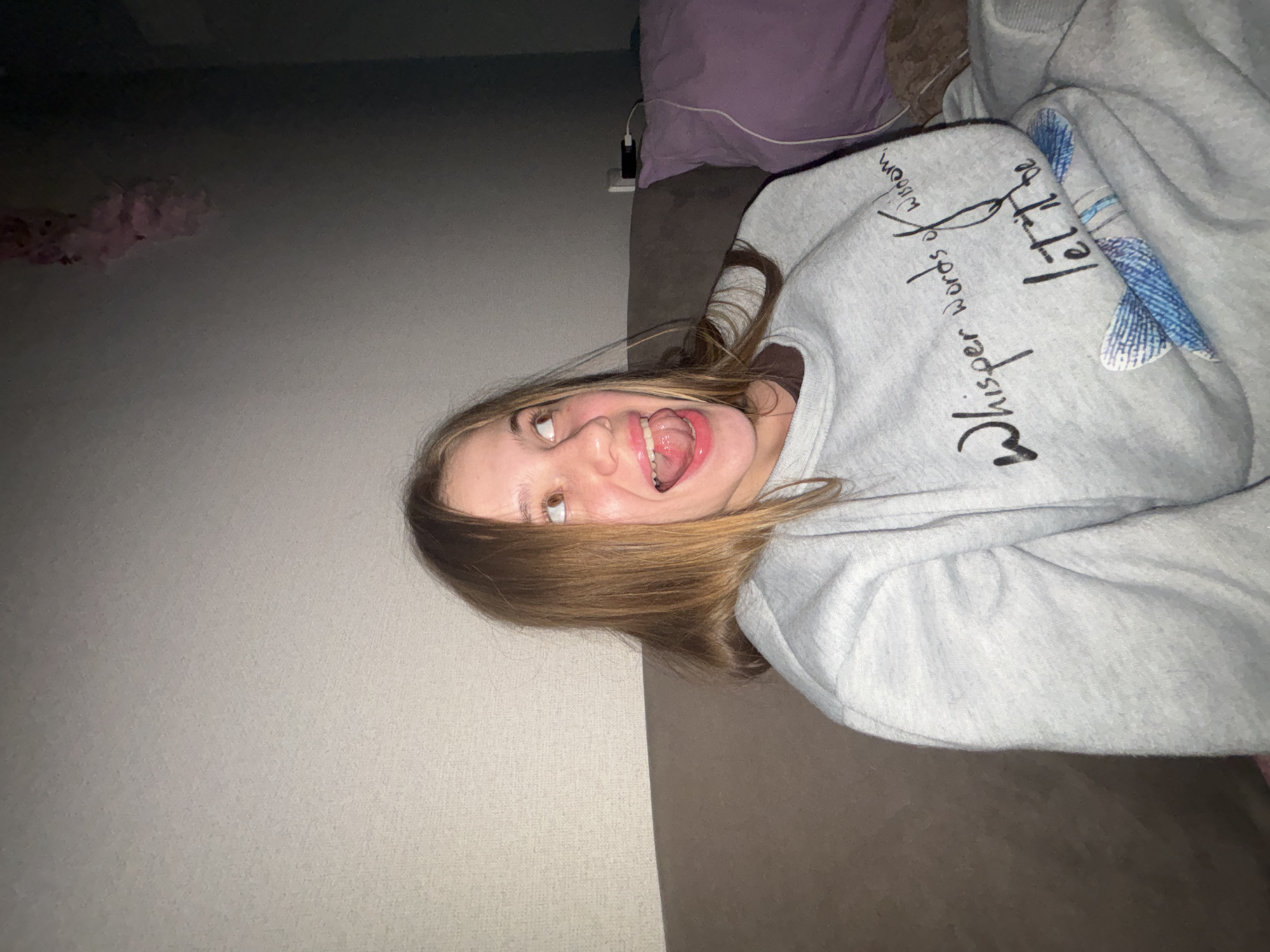 The image depicts a young Caucasian female with blonde hair, wearing a grey sweatshirt with the text "Whisper words of wisdom, let it be" and a blue fish design. She is seated on a brown sofa, with her mouth open and tongue sticking out, looking upwards and to her right. The background consists of a textured wall and a dark corner of the room, with a purple pillow and a power outlet visible. The lighting is dim, suggesting an indoor setting at night. The specific location within the city of Unknown, Russia, cannot be determined from the visual information provided.