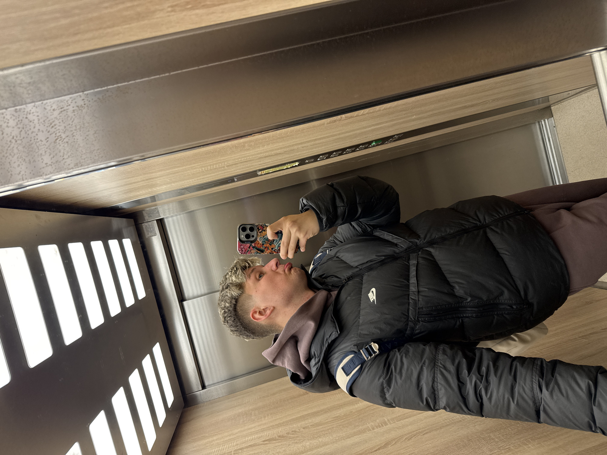 A young man stands in what appears to be an elevator, taking a selfie. He is wearing a black puffer jacket, a hooded sweatshirt, and holding a smartphone. The elevator walls are metallic with a wood-like paneling above. A light panel with a series of vertical, rectangular openings is visible on one side of the elevator. The location cannot be determined as Plovdiv, Bulgaria, from visual cues alone.