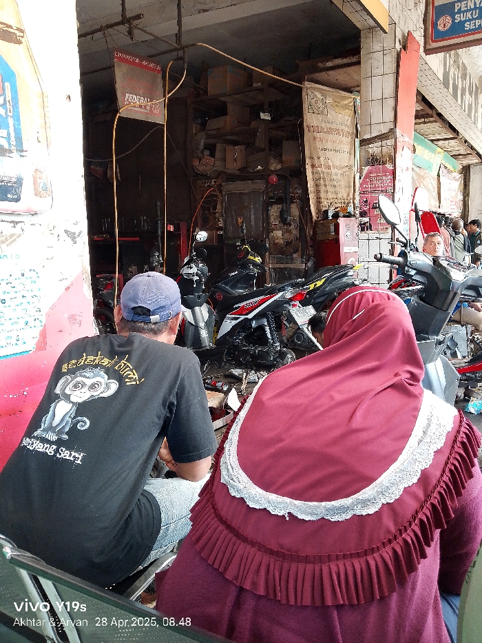 The scene takes place in a motorcycle repair shop in Kecamatan Haurgeulis, Indonesia. The shop is located in a densely populated area, with several buildings and a busy street. There are two people in the shop: a man wearing a black t-shirt with a monkey on it and a woman wearing a maroon headscarf with a white lace trim. The man is sitting on a stool, looking at a motorcycle. The woman is sitting on the floor, looking at the man. The shop appears to be a busy one, with several motorcycles parked around the shop and tools scattered about. The shop is messy, with papers and other debris scattered around. The shop is dimly lit, and the walls are painted in a dull, gray color. There are two motorcycles partially visible, one with a white and red color scheme and one with a black and red color scheme. The air inside the shop is thick with the scent of oil and gasoline.