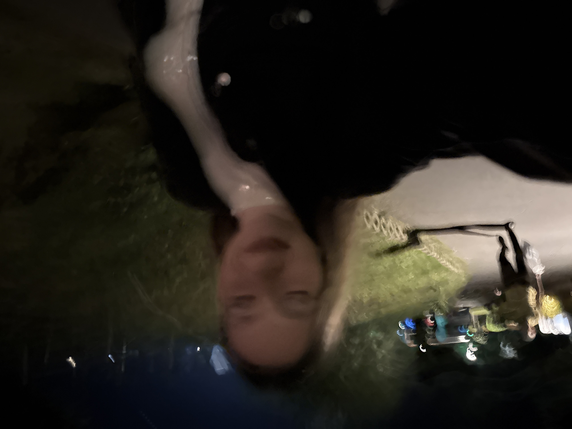 The image depicts a nighttime outdoor scene, characterized by significant motion blur and low light, likely taken in the city of Sozopol, Bulgaria.
In the immediate foreground, a female individual is captured from the chest up, positioned centrally. She has long, light-colored hair, and her eyes appear closed or heavily squinted due to blur. Her lips are slightly parted in what appears to be a smile. She is wearing a dark, possibly black, jacket or coat with visible buttons over a light-colored, high-collared garment. Her body is angled slightly to the left.
In the mid-ground, to the left of the primary subject, another individual is walking away from the camera on a paved pathway. This person is dressed in a light-colored, possibly olive or khaki, top and dark trousers. A light-colored object, potentially a bag, is visible in their left hand. Their figure is heavily blurred, indicating motion.
The environment surrounding the pathway consists of dark green vegetation, including bushes and potentially trees. A low, light-colored curb or barrier separates the path from the vegetation on the left. In the background, numerous artificial light sources are visible as blurred orbs of varying intensity and color, including white, blue, red, and green. These lights suggest the presence of nearby structures, entertainment facilities, or illuminated landscaping. The overall scene is dark, indicative of evening or nighttime conditions.
