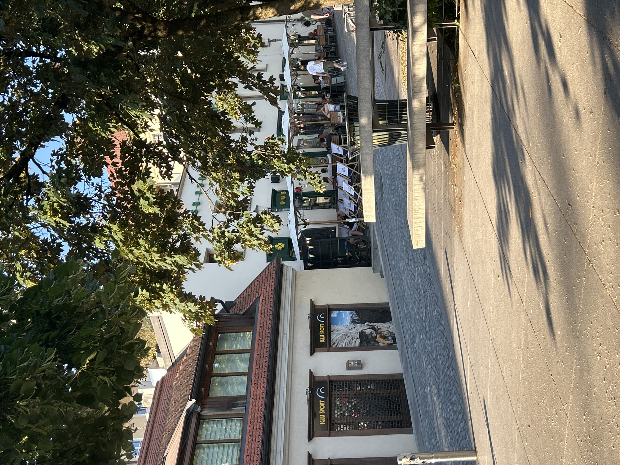 An outdoor scene captures a European-style street with buildings, a paved walkway, and a seated area. The buildings have traditional architecture, with terracotta roofs and cream-colored facades. One building houses two shops with the "IGLU SPORT" logo above their entrances. The shop to the right displays a graphic of a climber on a rock face.
In front of the buildings, a cobbled street slopes upwards. Adjacent to the street is a paved area with outdoor seating, shaded by umbrellas. Several people are seated at tables, and some lounge chairs are arranged near the umbrellas. A man in a white t-shirt and khaki shorts is walking away from the viewer towards the seating area. Another person is cycling in the background.
The foreground features a large concrete bench with metal legs, casting a shadow on the paved ground. The ground is a mix of large square tiles and smaller cobblestones. Sunlight casts long shadows across the scene, indicating it is likely late afternoon or early morning. Trees with green foliage overhang the scene, dappling the ground with shade and light. The sky above is clear and blue.