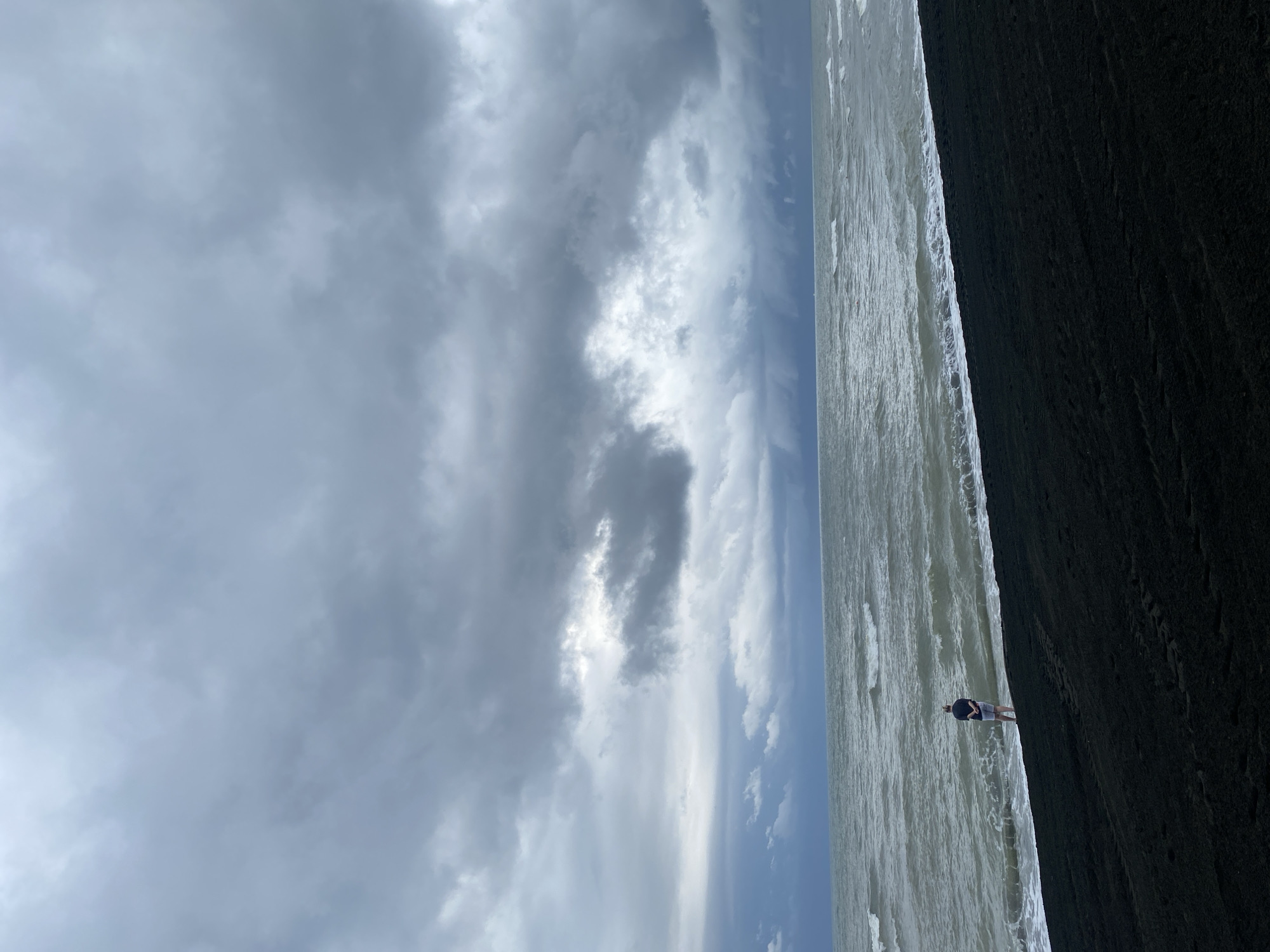 In Ureki, Georgia, on a black sand beach, a person stands near the water's edge under a dramatic, cloudy sky. The person, viewed from behind and from a distance, is wearing a dark shirt and light shorts. They are facing the turbulent, grey ocean with waves crashing onto the shore. The black sand covers the foreground, with subtle tracks visible. The horizon is dominated by heavy, dark clouds, suggesting impending rain or a storm. The overall atmosphere is somber and natural, capturing a moment of solitude against a powerful natural backdrop.