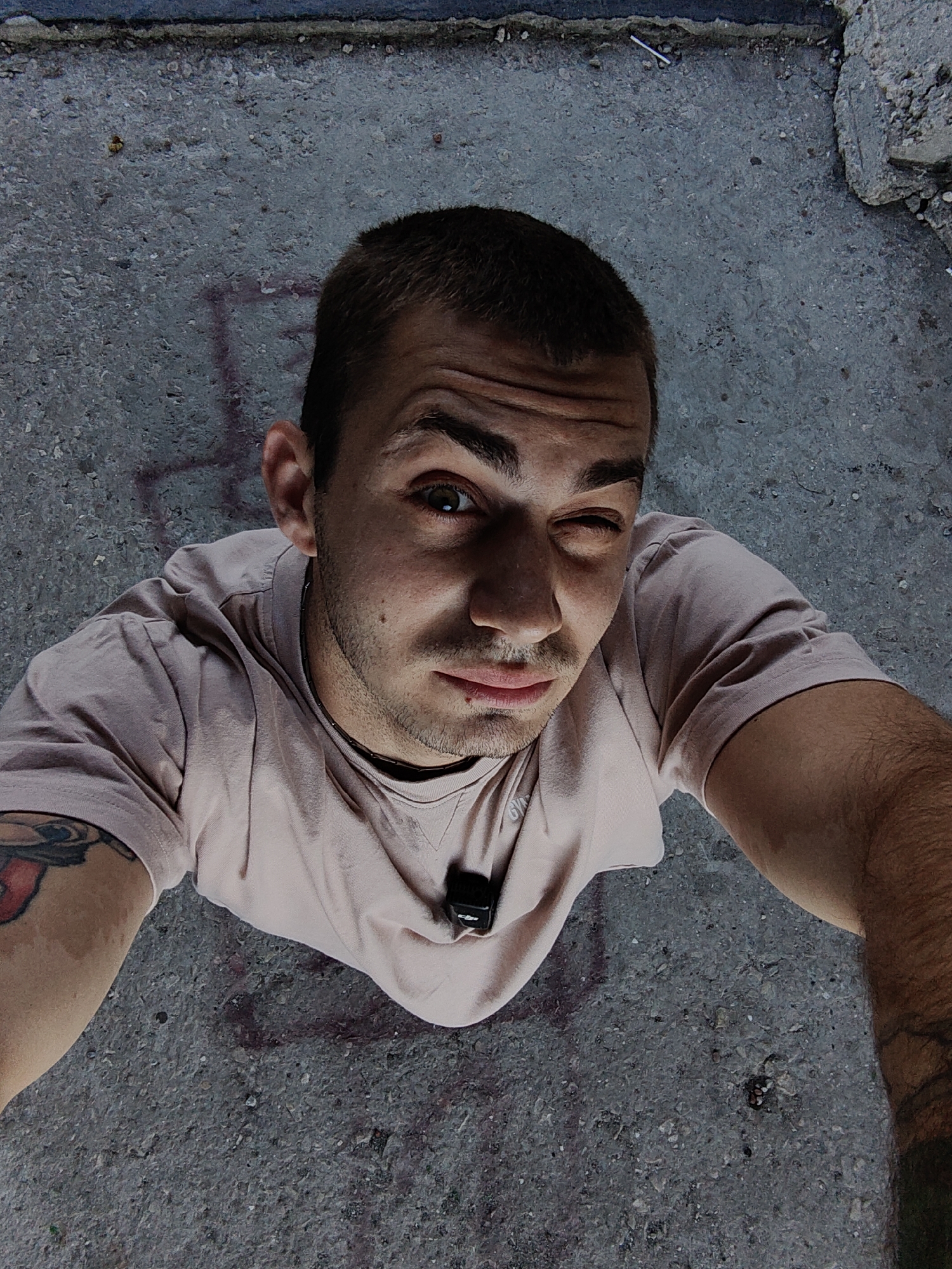An overhead, medium shot captures a young adult male with short, dark hair and a light tan t-shirt looking up at the camera. He is outdoors on a rough, gray, concrete surface, possibly an urban street or sidewalk, with some debris and faint red markings visible. The man is winking with his left eye, his right eye open and looking directly at the camera. He has stubble on his face and a small mark or cut on his lower lip. A black clip-on device is attached to his t-shirt, near his collarbone. A tattoo is partially visible on his left arm. The lighting suggests it is daytime, with shadows cast on the ground. The environment does not provide specific indicators of Varna, Bulgaria.