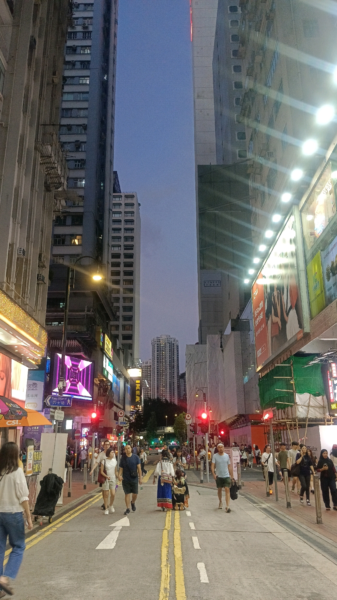 The vertical, high-angle full shot depicts a street scene in Hong Kong at dusk. Towering, modern buildings line both sides of the street, their facades adorned with numerous windows and illuminated signs. A woman in a white blouse and blue jeans walks away from the camera on the left sidewalk. On the street, a group of people walk towards the camera. A couple holds hands in the center, the woman in a white top and shorts, the man in a navy blue t-shirt and shorts. To their right, a person in traditional clothing walks with a smaller person in similar attire. More pedestrians are visible further down the street, with streetlights casting a warm glow. Traffic lights display red signals. The sky above is a deep twilight blue.