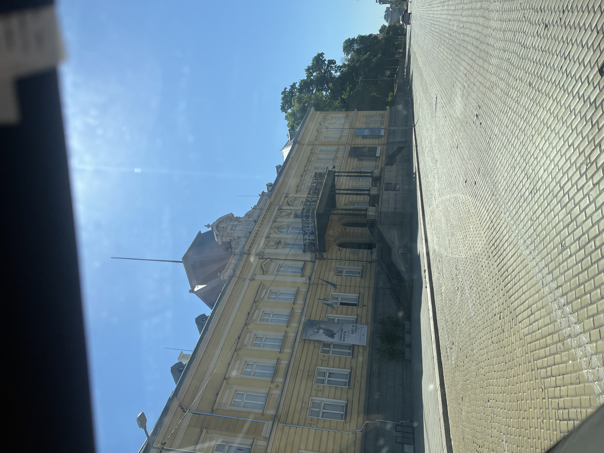 An exterior, eye-level shot captures a yellow, historical building on a clear, sunny day. The building, likely a museum or public institution, features a grand facade with numerous windows and an ornate balcony. A banner with Cyrillic text and an image of a bird is prominently displayed on the building's lower level. Two flags, one with horizontal stripes of white, green, and red, and another blue flag, hang near the entrance. The building is situated on a wide, cobblestone street, with lush green trees lining the right side of the road. A few cars are visible in the distance, suggesting an urban environment. The sky is a vibrant blue, with faint streaks of sunlight creating a lens flare effect in the upper portion of the image.