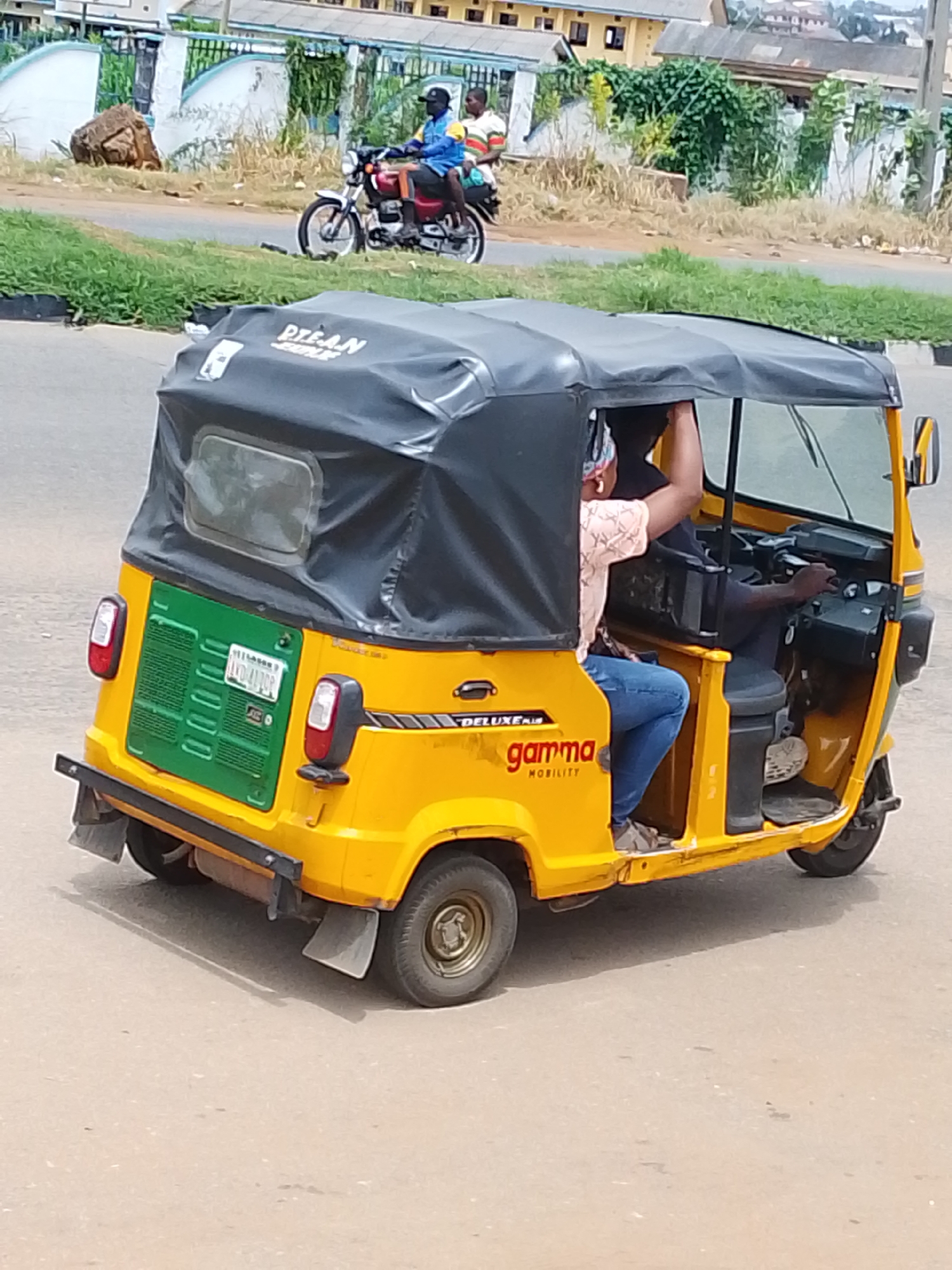 A yellow motorized rickshaw, also known as an "Okada" or "Keke Napep", is captured from a rear-right, slightly elevated angle on a paved road. The rickshaw features a black canvas roof and a green rear panel with a license plate reading "AGD 4170". A person, wearing a pink patterned shirt and blue jeans, is seated in the passenger area. The driver, visible from the side, is wearing a dark shirt and appears to be operating the vehicle. In the background, a red and white motorcycle with two occupants is visible on the road. Further back, buildings and lush green vegetation line the street, suggesting an urban or semi-urban setting. The overall scene depicts a moment of transportation and daily life in what is identified as Oja Oba, Oko Erin, Nigeria.