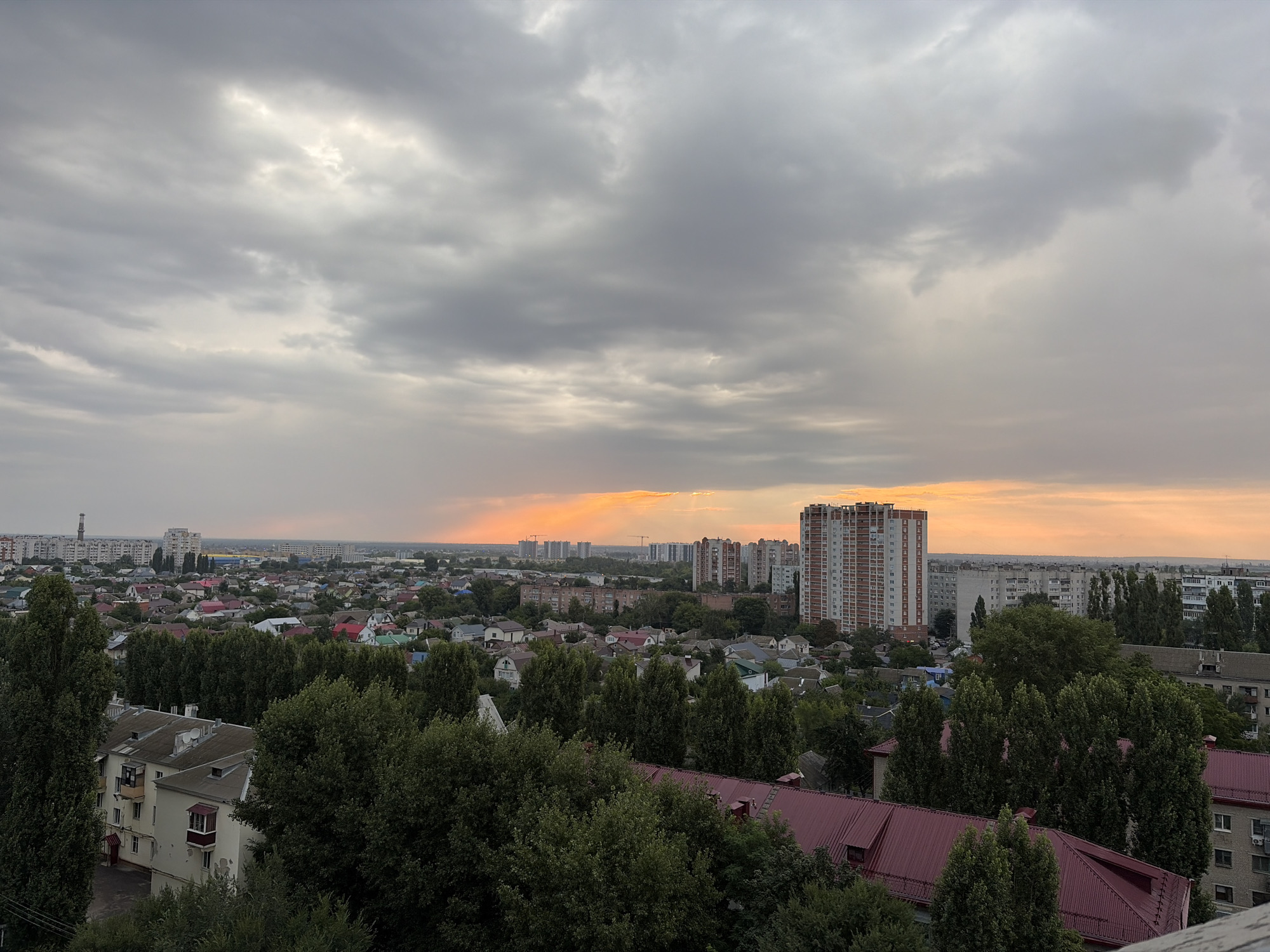 This is a wide shot of the city of Novaya Usman, Russia, at dusk. The sky is overcast with dramatic clouds, and a sliver of orange light from the setting sun is visible on the horizon, peeking through a break in the clouds. The foreground is dominated by the dark green foliage of trees, with the red roofs of houses visible beneath them. In the mid-ground, a dense residential area with various sized buildings, including apartment complexes and smaller houses with colorful roofs, is spread out. The background features a cityscape with taller apartment buildings and industrial structures. The overall impression is of a densely populated urban area under a moody, darkening sky.