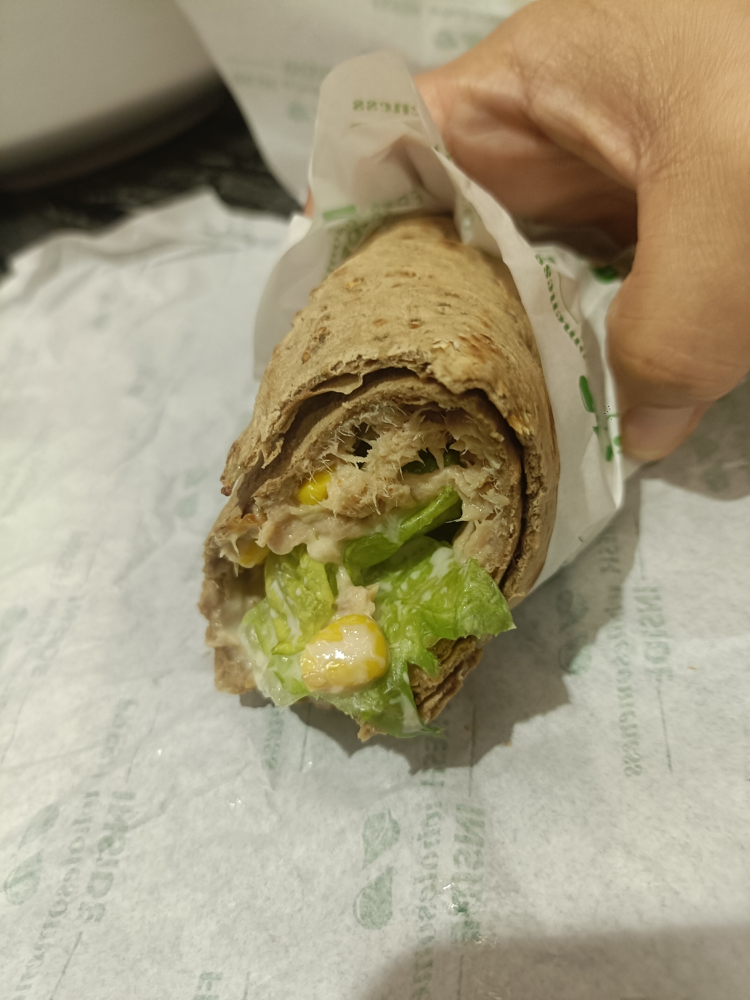 A close-up shot shows a whole wheat wrap with visible fillings of lettuce, corn kernels, and a creamy sauce, possibly mayonnaise or a salad dressing. The wrap is held by a person's hand, which is partially visible on the right side of the frame. The wrap is placed on a white paper wrapper, which has green text printed on it. The background is blurred, with a hint of a dark surface on the left and a white, possibly food-related, surface above. The lighting suggests an indoor setting. The image does not contain information that would allow for the identification of its location in Dubai, United Arab Emirates.