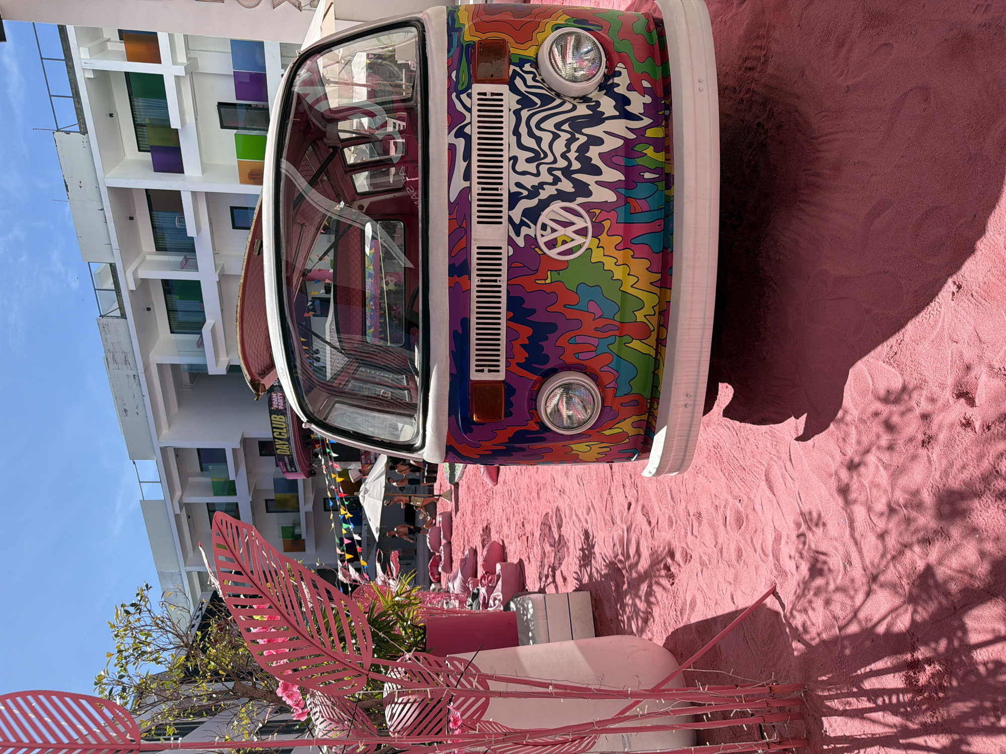 The image shows a vibrantly decorated Volkswagen bus parked on pink sand, with a modern building in the background. The bus is adorned with a psychedelic, multi-colored paint job featuring wavy patterns in shades of blue, purple, green, yellow, and orange. The Volkswagen logo is prominently displayed on the front. The scene is bathed in bright sunlight, casting strong shadows on the sand, suggesting it is daytime.
In the background, a multi-story white building with colorful square windows in shades of orange, blue, green, and purple can be seen. In front of the building and surrounding the bus, there are various pink decorative elements, including large leaf-shaped metal cutouts and string lights. Some triangular flags are also visible, adding to a festive atmosphere.
A sign on the side of the bus reads "DAY CLUB FUN PARTY," indicating a potential venue for entertainment or leisure. Although no people are clearly visible, the setting suggests a lively outdoor establishment, possibly a beach club or a themed event space. The overall impression is one of a fun, energetic, and visually striking environment. The location context indicates this is likely Baan Bang Rak, Thailand.