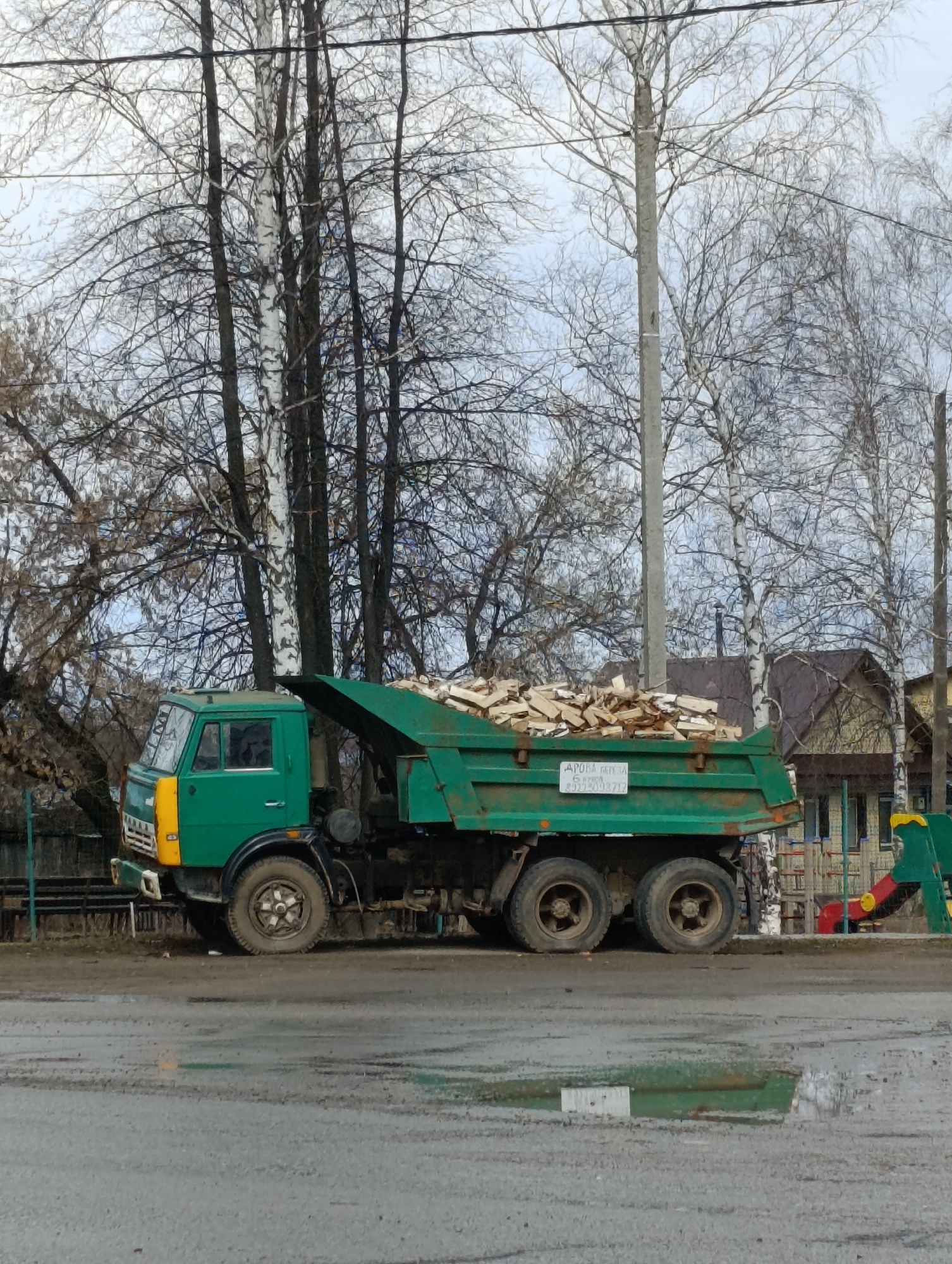 The image shows a green dump truck loaded with what appears to be lumber. The truck is parked on a wet, possibly muddy, road with puddles visible. The cab of the truck is green with a yellow vertical stripe on the front. The truck's bed is raised slightly. Behind the truck are trees, some with white bark, and a building that appears to be a house or small structure. A playground structure with a slide is also visible. The sky is overcast. The lettering on the truck, though partially illegible, shows Russian characters. The location is Chaikovsky, Russia. The general atmosphere appears to be urban or suburban, possibly a residential area. There are no visible people.
