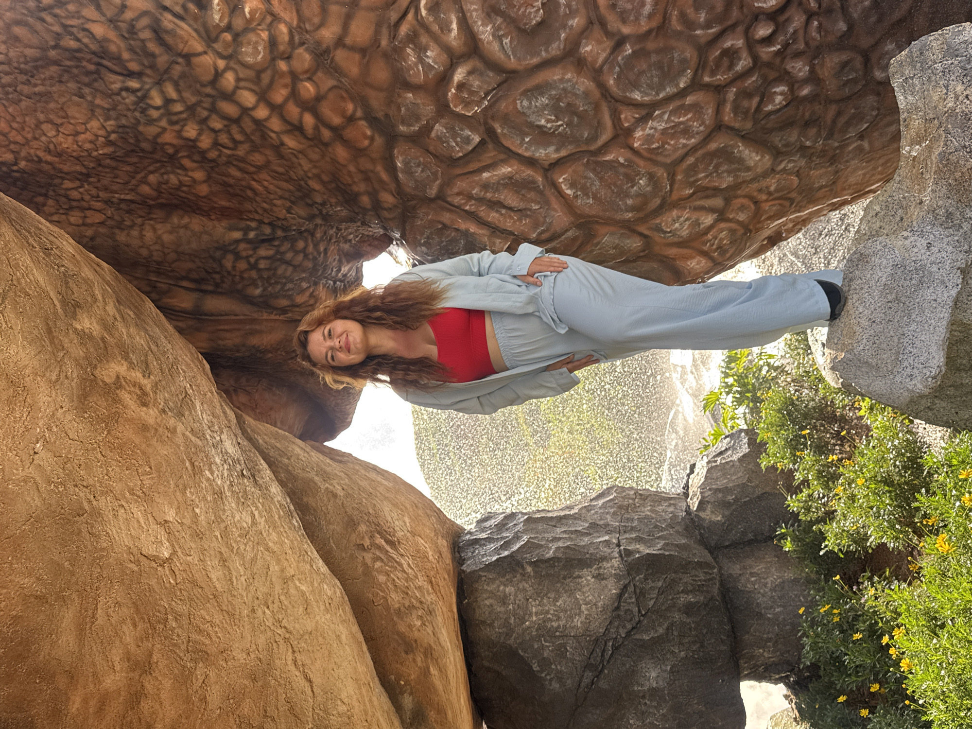 A woman stands in a rocky, naturalistic enclosure with a faux reptile skin texture on the right. She is wearing a red bandeau top and light blue wide-leg pants and an open jacket. She is standing on a rock formation. Behind her, there is a misty water feature and green foliage with yellow flowers. The overall setting appears to be an outdoor attraction. The image was taken in Thôn 1, Vietnam.