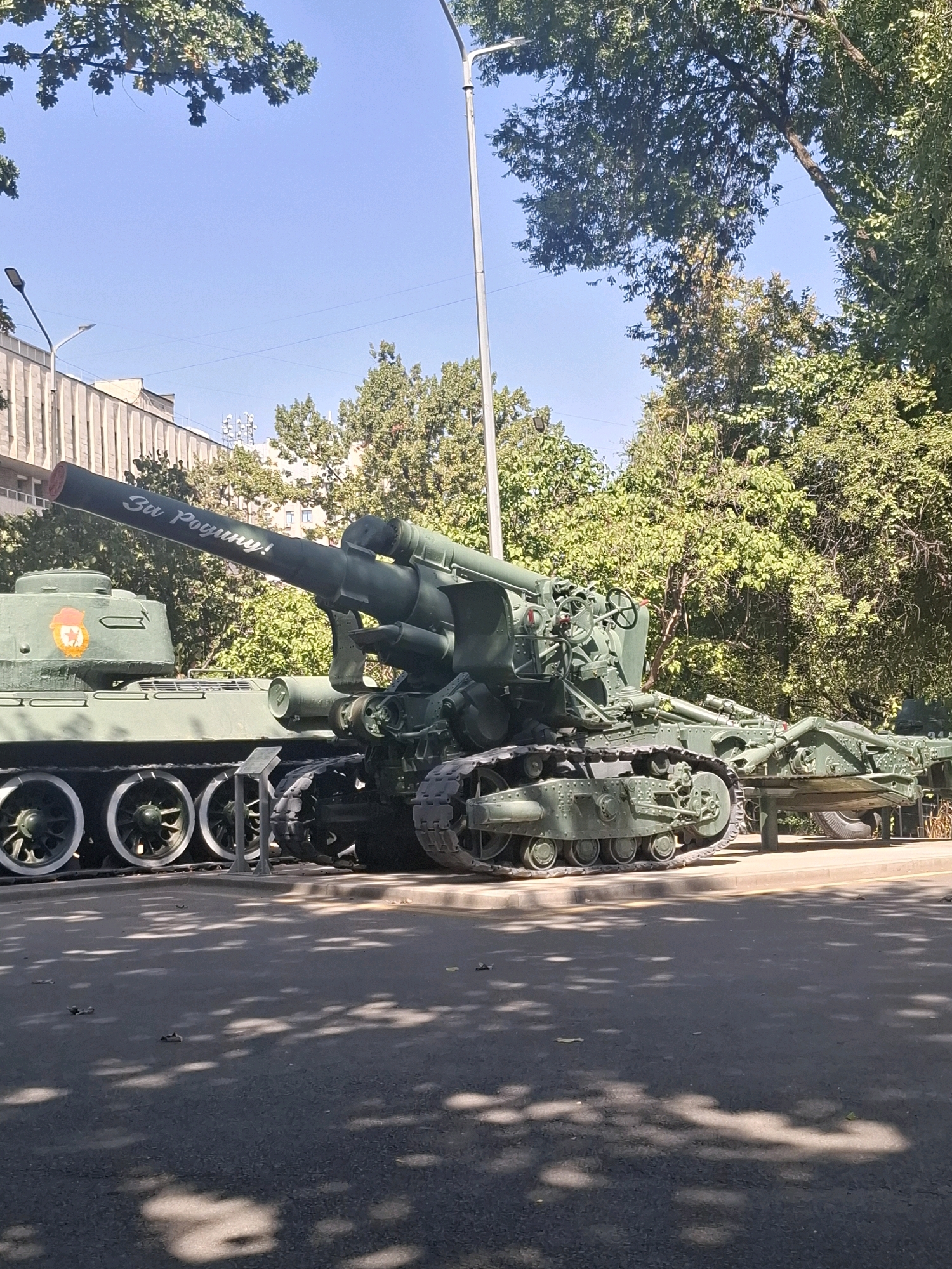 The image depicts a military exhibition featuring a green tank and a large artillery piece, both in a state of disrepair or preservation, likely as a monument. The artillery piece has the inscription "За Победу!" ("For Victory!") on its barrel. Behind the military equipment, a building with light-colored vertical elements is visible, along with trees and a clear blue sky. The ground in front of the exhibit is paved, with shadows from trees cast across it, suggesting an outdoor public space. The presence of streetlights and overhead wires indicates an urban setting. Based on the inscription and the context of military monuments, this scene is located in Almaty, Kazakhstan, specifically at the "Eternal Flame" memorial complex in Panfilov Park, which commemorates World War II soldiers. No people are visible in the frame.