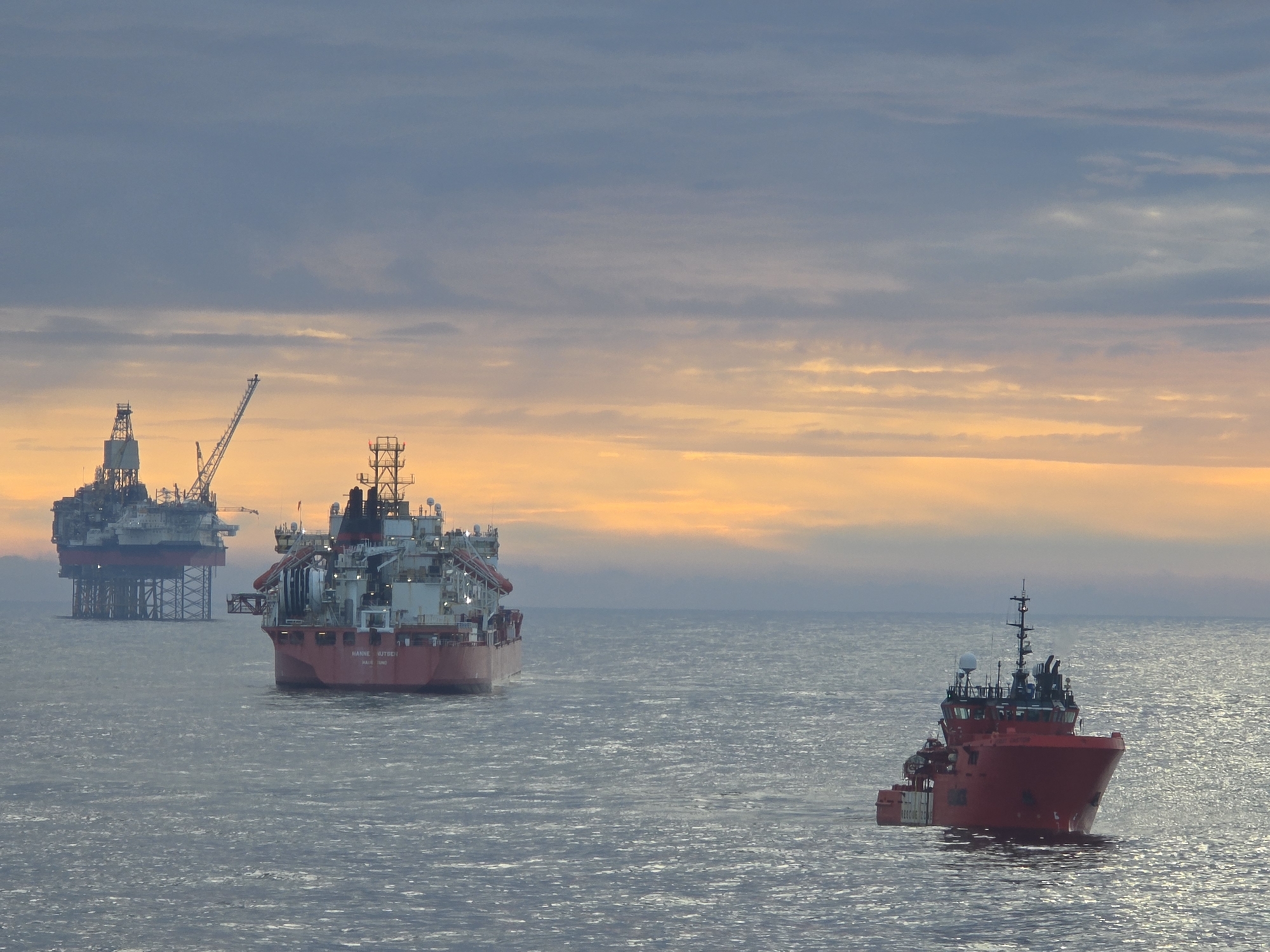 The image depicts a marine environment at dusk or dawn with an orange-tinted sky. In the background, there is an oil rig platform. A large supply vessel is positioned closer to the viewer, facing away. A smaller, red vessel is in the foreground, also facing the viewer. The water is calm with subtle ripples. Location: Unknown, Unknown.