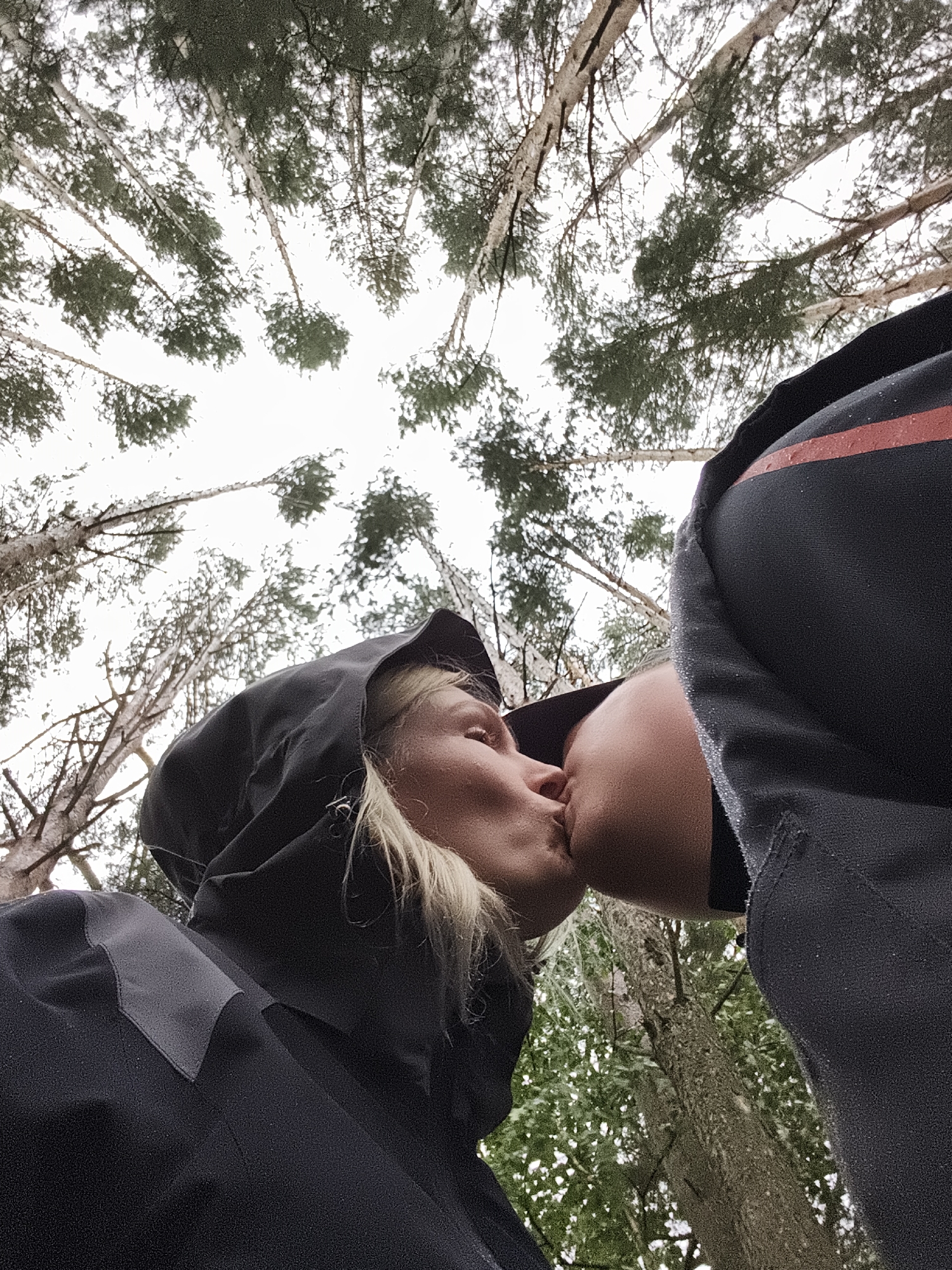 In this low-angle shot, two people are captured in an intimate moment, sharing a kiss amidst a forest setting. The viewpoint looks upwards towards the canopy of tall trees, with their branches reaching towards a bright, overcast sky. The foliage suggests a temperate climate, possibly during spring or summer.
One subject, a woman with blonde hair, is wearing a black hooded jacket. Her face is turned towards the other person, eyes closed, and lips puckered in a kiss. The other subject is only partially visible, with their face obscured by the embrace. They are also wearing dark clothing, with a distinctive red stripe visible on their shoulder area. The texture of the clothing suggests a water-resistant material, possibly hinting at damp conditions or recent rain, as small droplets are visible on the fabric.
The overall mood is romantic and peaceful, amplified by the natural backdrop of the forest. The time of day appears to be daytime, given the brightness of the sky, though the overcast conditions create a diffused light. The setting is clearly outdoors, within a wooded area. There is no visible text in the image. The location context provided is Русский Вожой, Russia.