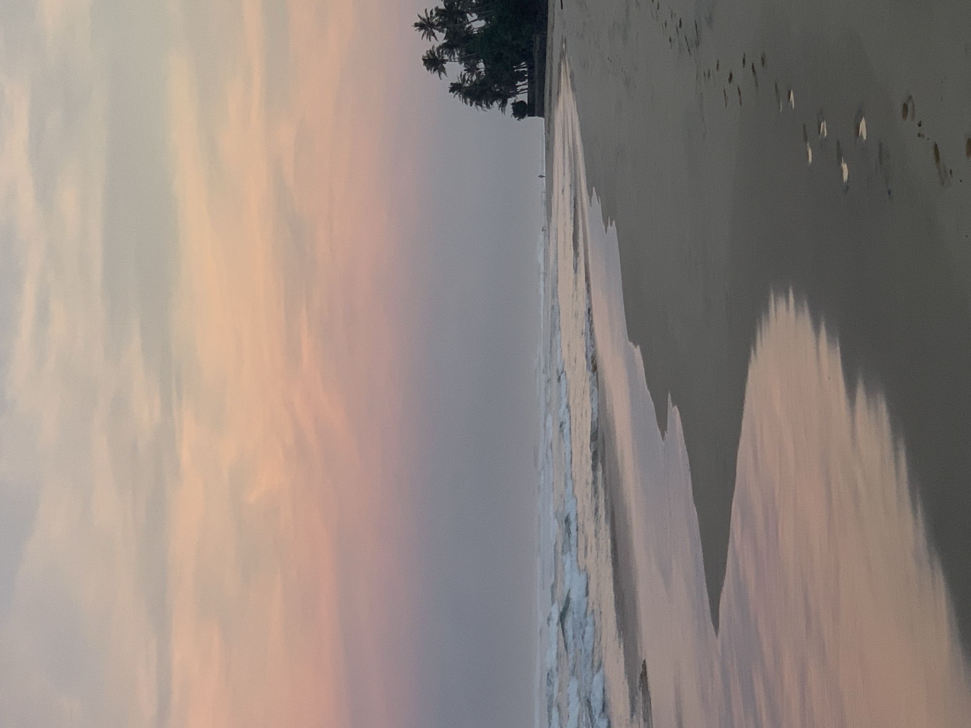 The image depicts a beach scene in Kathaluwa, Sri Lanka. The sky is filled with pink and grey clouds, indicating either sunrise or sunset. The ocean meets the beach, with visible waves and foam. On the right side, the sandy beach stretches into the distance, showing footprints. In the midground, there is a line of palm trees. The scene appears calm and serene.