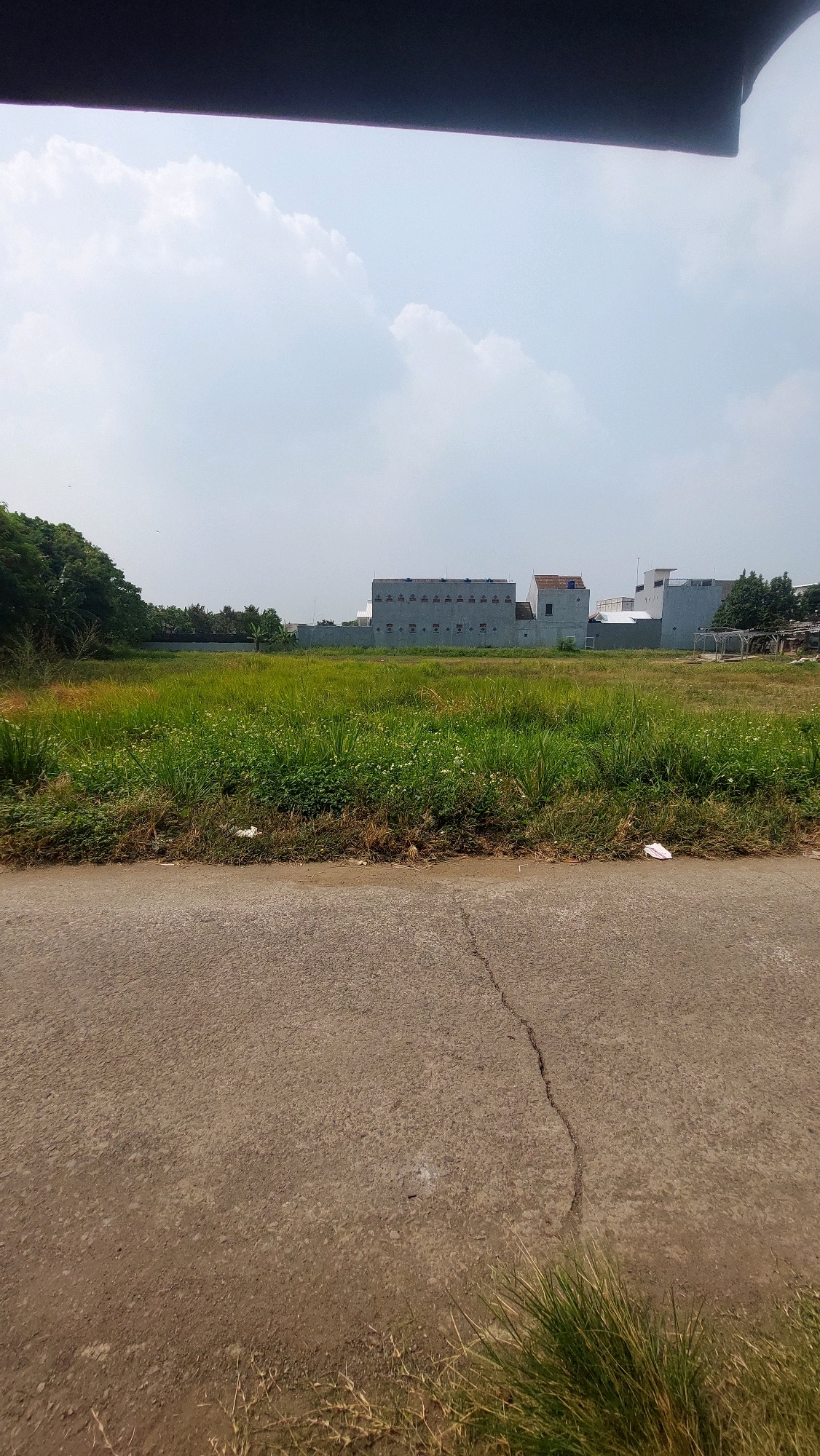 An outdoor, eye-level, vertical shot captures a vacant lot in Bojong Gede, Indonesia. In the foreground, a cracked asphalt road runs horizontally across the bottom of the frame. Beyond the road lies a field of green grass, dotted with small white flowers, extending towards a line of trees on the left. In the middle ground, a long, gray building with a repetitive pattern of small, dark rectangles is visible, flanked by other structures and a wall. The sky above is a pale blue, filled with scattered white clouds, and a dark, indistinct object is visible at the top of the frame, possibly a roof overhang or awning. The scene appears to be undeveloped and potentially part of a construction site or an area awaiting development.