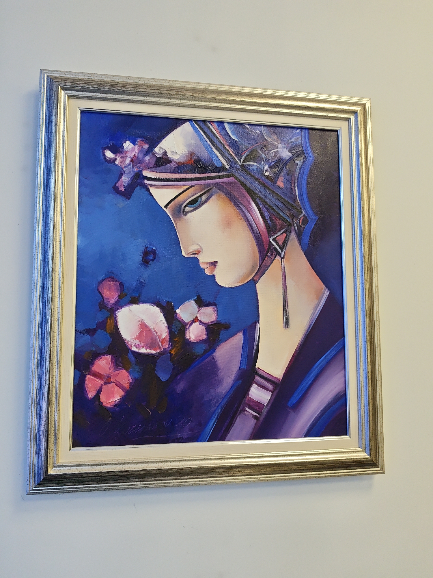 The image shows a framed painting of a woman in profile wearing a headdress. The painting features a vibrant blue background with pink and purple flowers. The woman has a stylized face with prominent eyes and a slender nose. She is wearing a purple garment with blue accents. The frame of the painting is silver and gold with a textured finish. The painting is hanging on a plain, light-colored wall. This painting is located in the Pleven Regional Historical Museum in Pleven, Bulgaria.