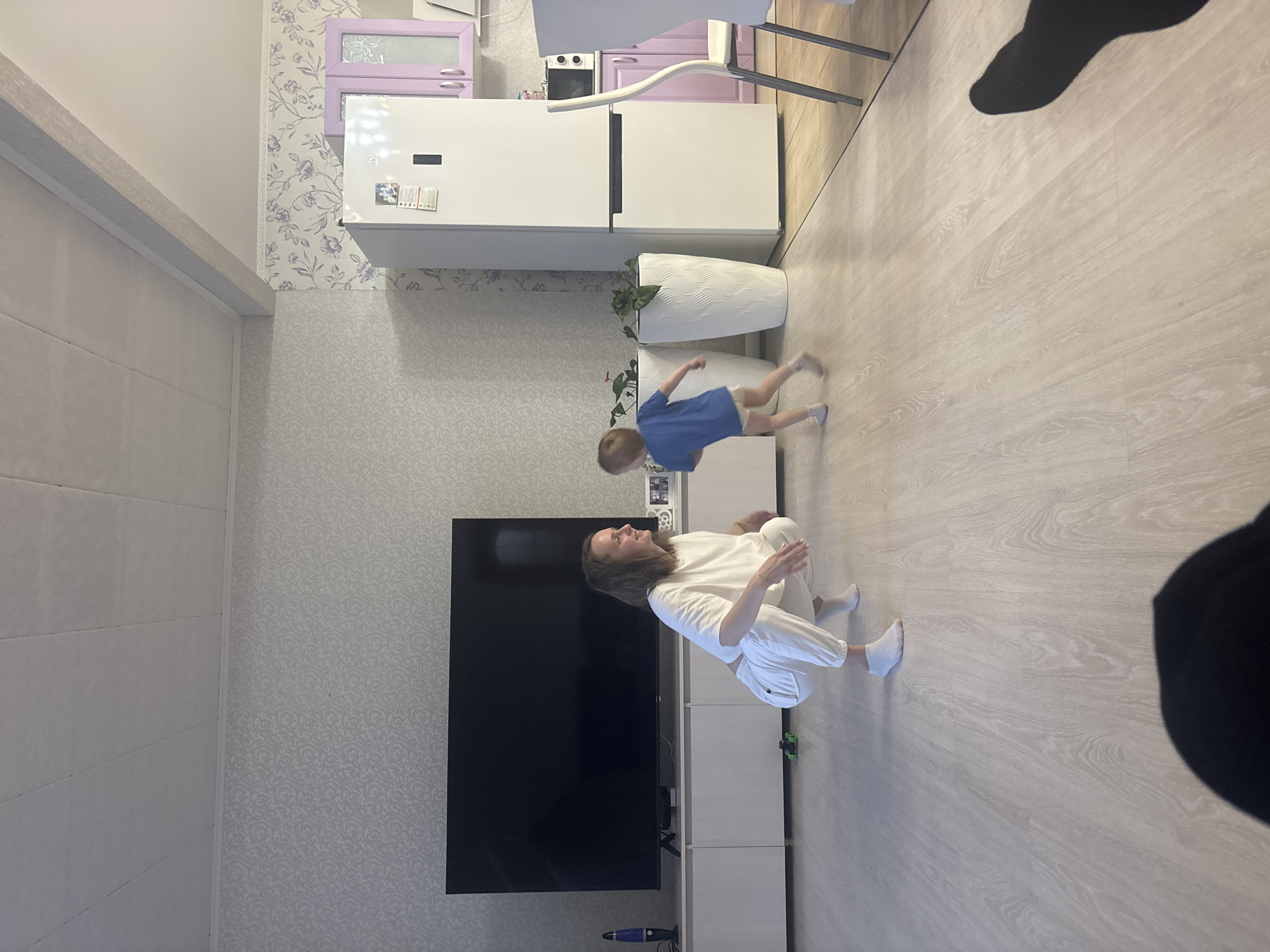 The image depicts the interior of a living space in Nestiukovo, Russia. A woman is sitting on the floor wearing a white top and white pants. A young child wearing a blue shirt and beige shorts is running near her. A large black television is mounted on the wall. A white refrigerator is visible in the background, along with kitchen cabinets in lavender and white. Two white planters are present next to the refrigerator. The floor appears to be light-colored wood laminate. There is patterned wallpaper on the walls. A vacuum cleaner is visible near the TV.